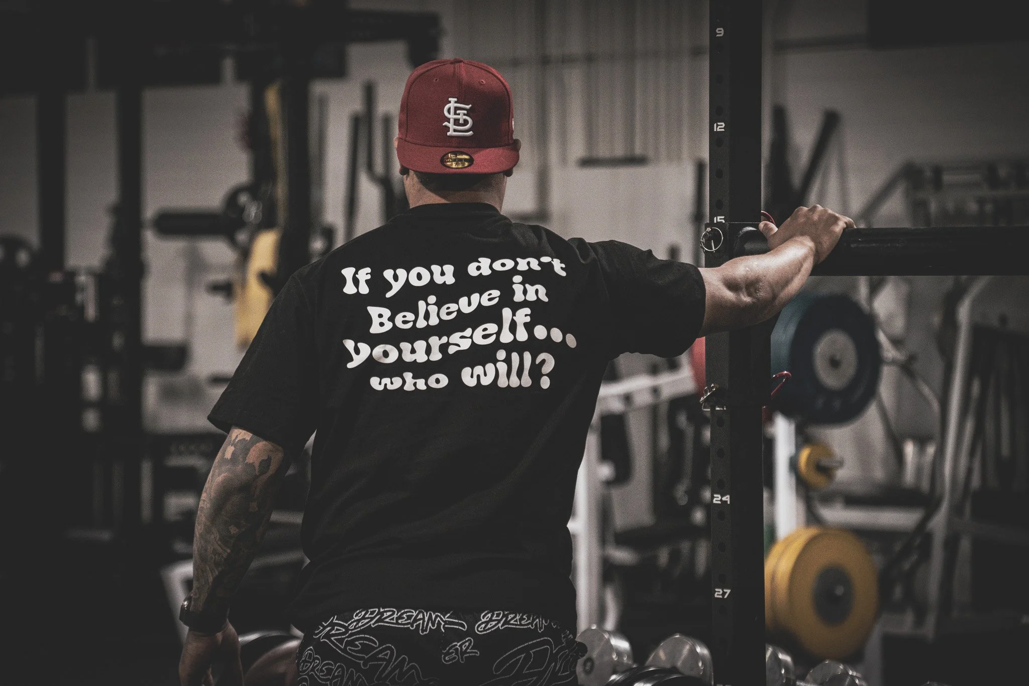 A man in a red baseball cap and black t-shirt with motivational text stands in a gym, leaning on a squat rack with weights in the background.