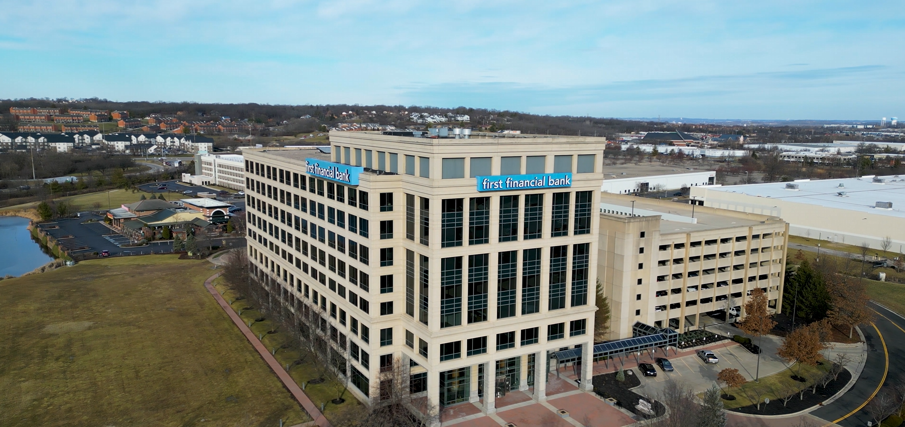 Aerial view of a beige office building with blue signs that read 'first financial bank' on the top floors. The building is surrounded by parking lots, trees, and nearby industrial and residential areas under a partly cloudy sky.