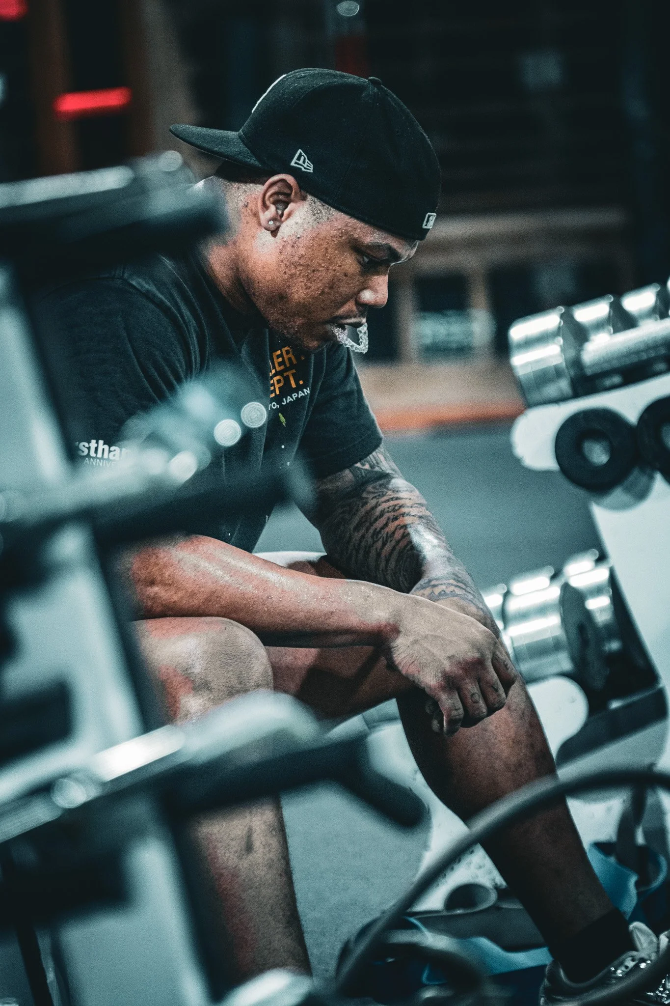 A young man with tattoos, wearing a black baseball cap backwards and a black T-shirt, is sitting on a motorcycle, resting his arms on his knees and looking down thoughtfully.