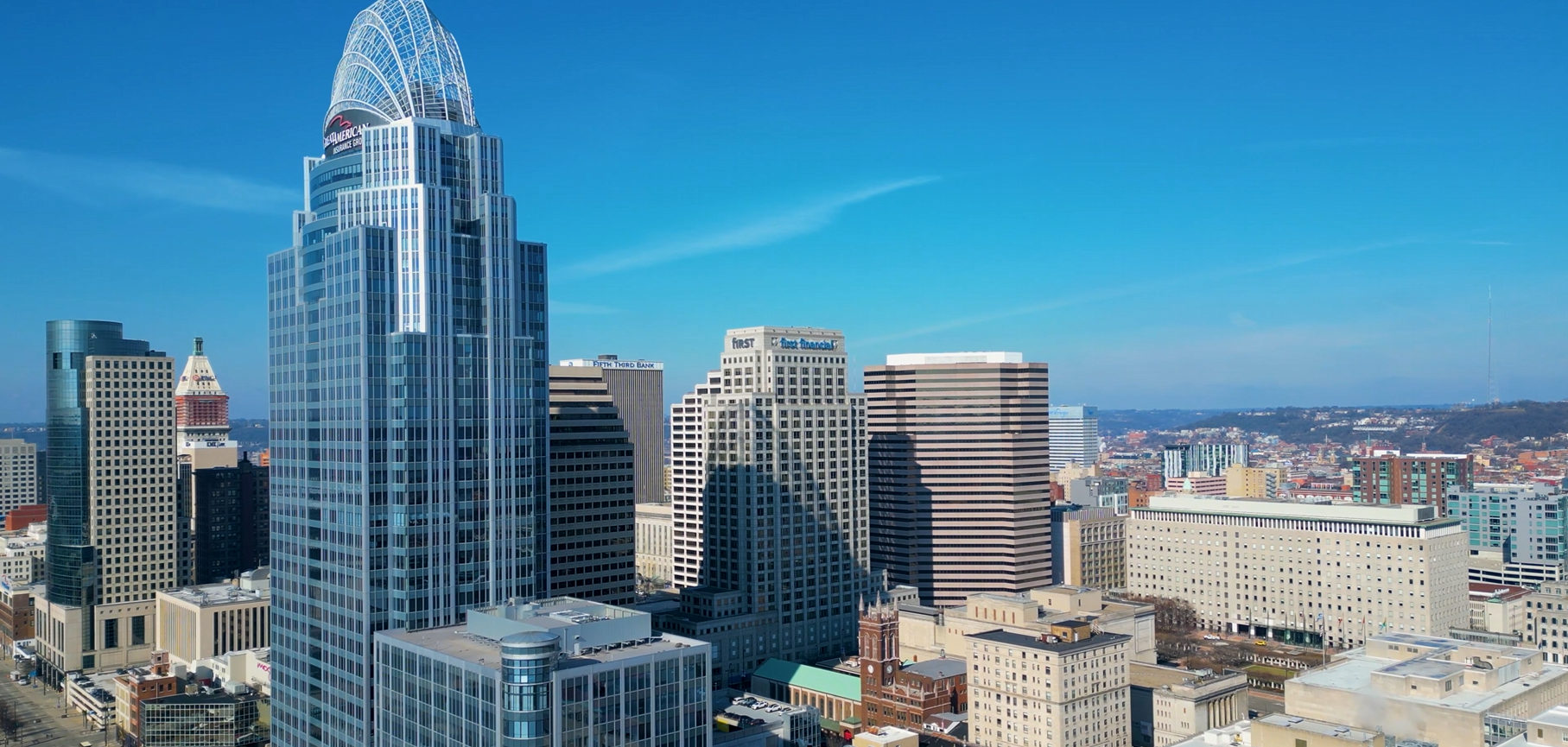 A city skyline featuring tall skyscrapers, including a prominent glass high-rise with a curved top, under a clear blue sky.