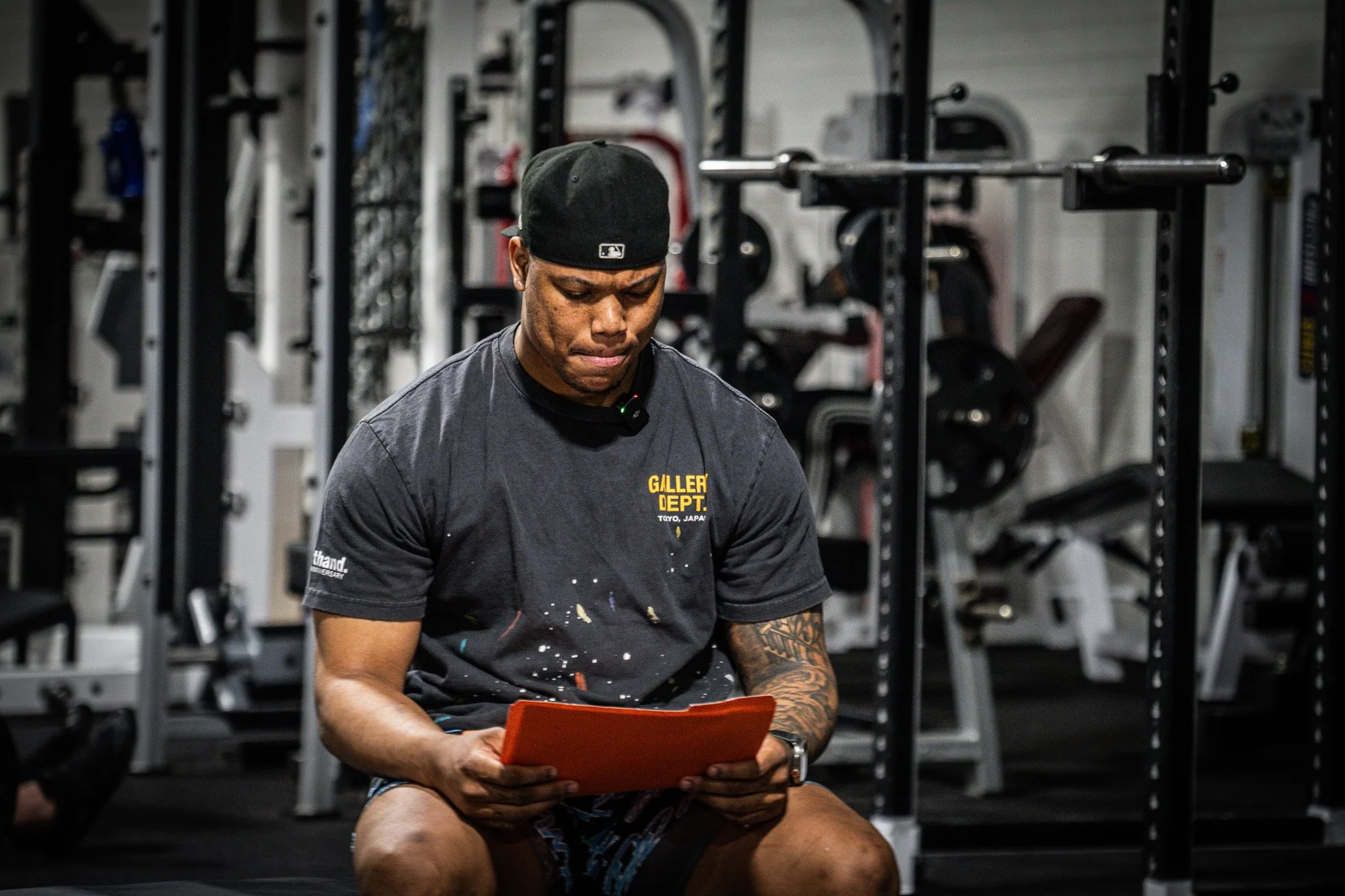 A man sitting in a gym, looking at an orange tablet, wearing a black cap and a black t-shirt with paint splatters, in front of exercise equipment.