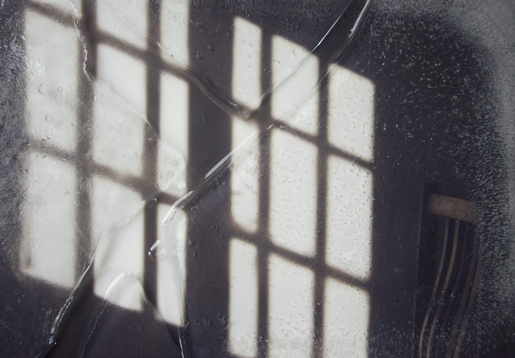 Reflexo de grades de uma janela em uma superfície molhada, com gotas de água e sombra do ambiente ao redor.