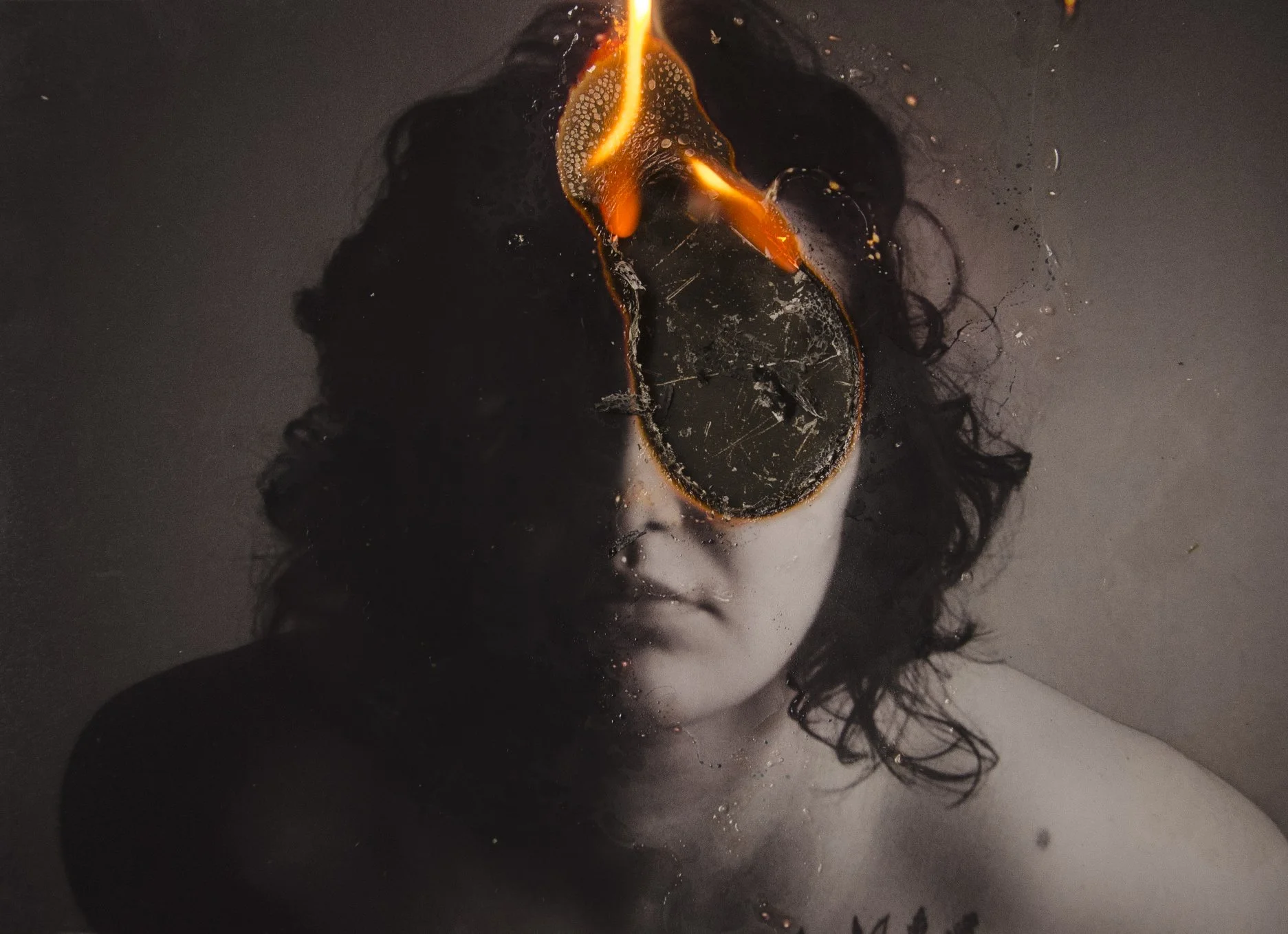 Pessoa com cabelo cacheado, parcialmente submersa em água escura, com uma poça de líquido escuro no rosto, cobrindo os olhos, que parece estar queimando ou derretendo.