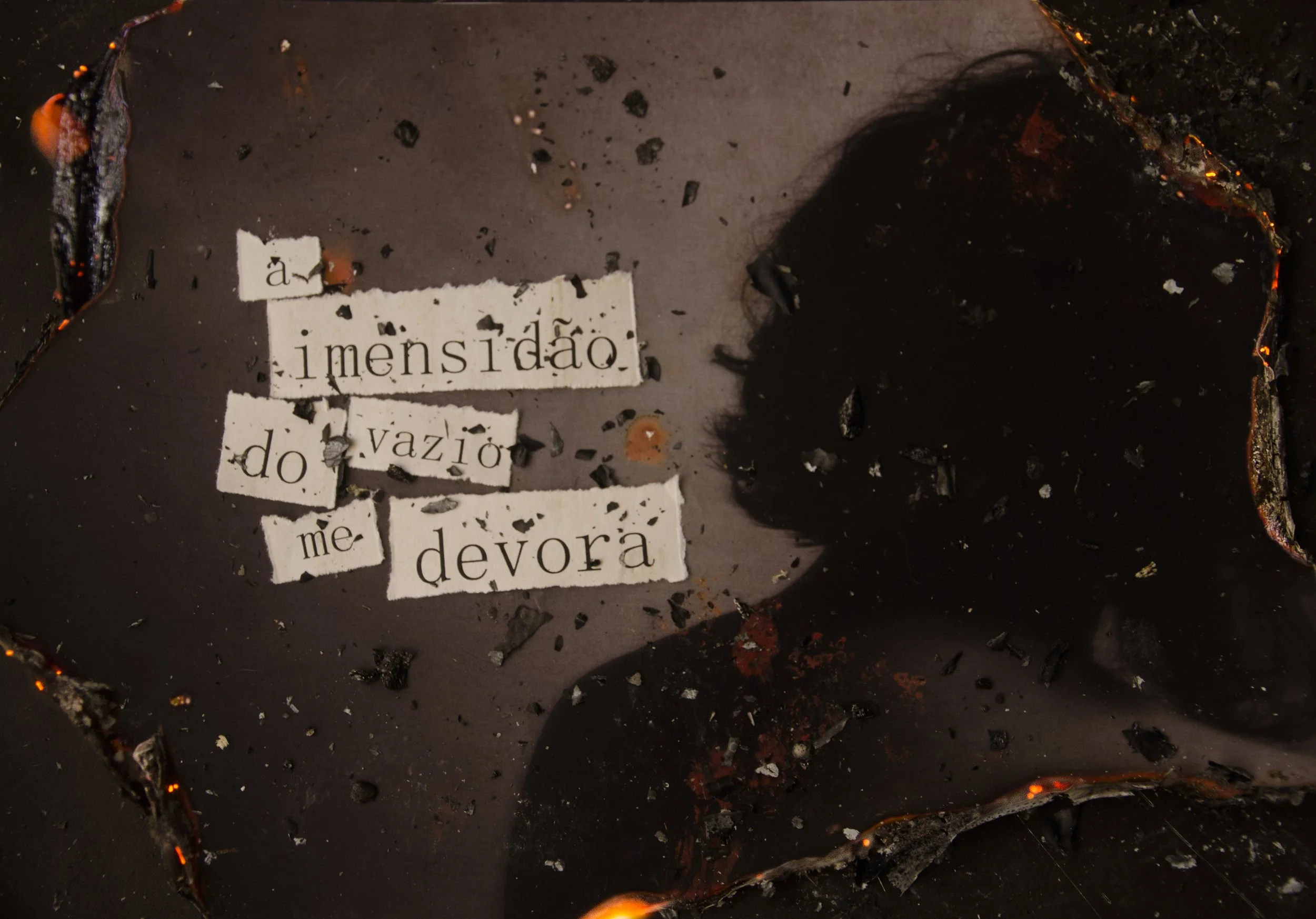 Pedaços de papel manuscrito com a frase 'a humanidade do vazio me devora', cercados por destroços e fragmentos de material escuro, possivelmente metal ou carvão, em uma superfície escura e queimada.
