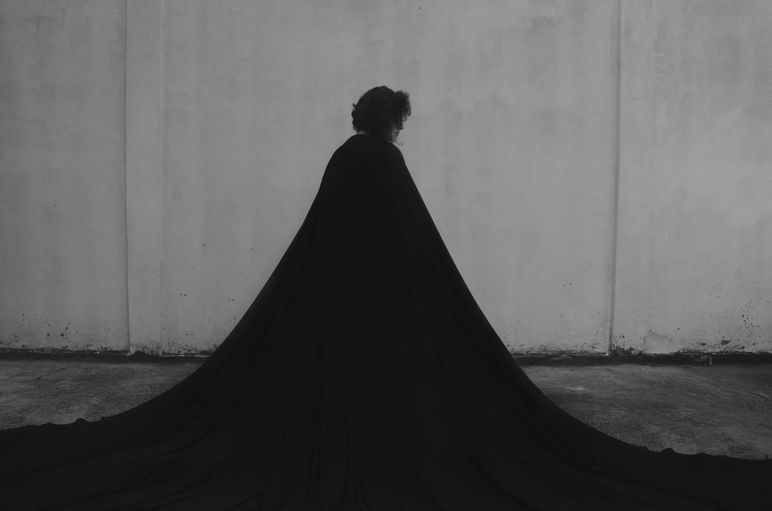 Pessoa com cabelos cacheados usando um vestido preto longo que se arrasta pelo chão, de lado, em frente a uma parede cinza.