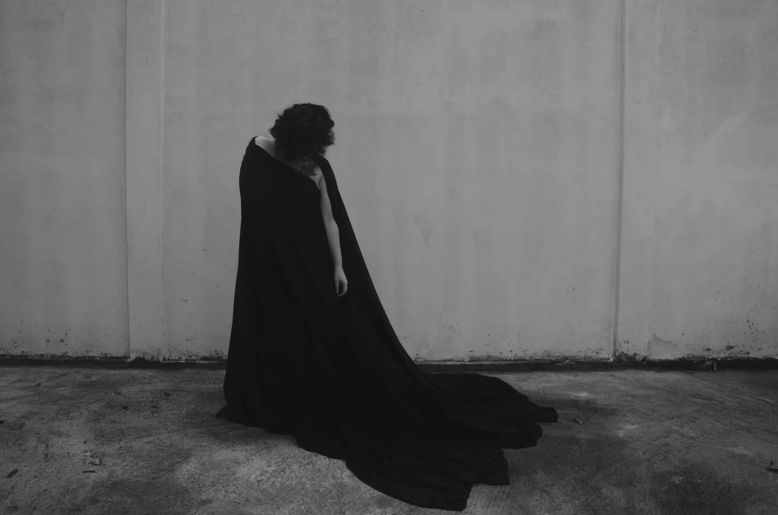 Pessoa com cabelo encaracolado usando vestido longo preto, solta no chão, em um espaço com parede de concreto, na cor preta e branca.
