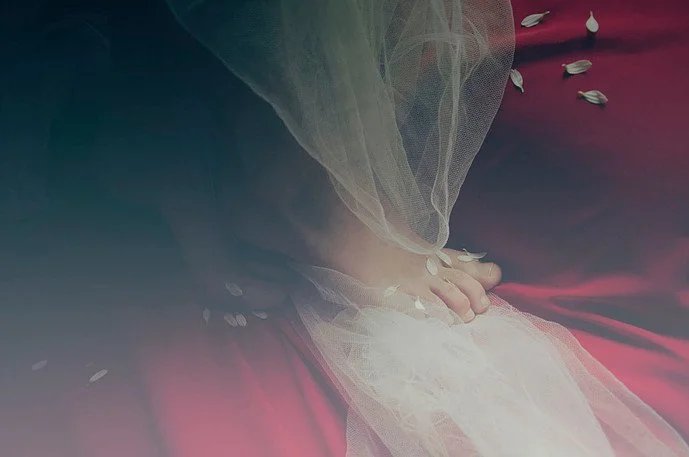 Pé de uma mulher parcialmente coberto por um véu, sobre um tecido vermelho coberto por pétalas de margaridas brancas