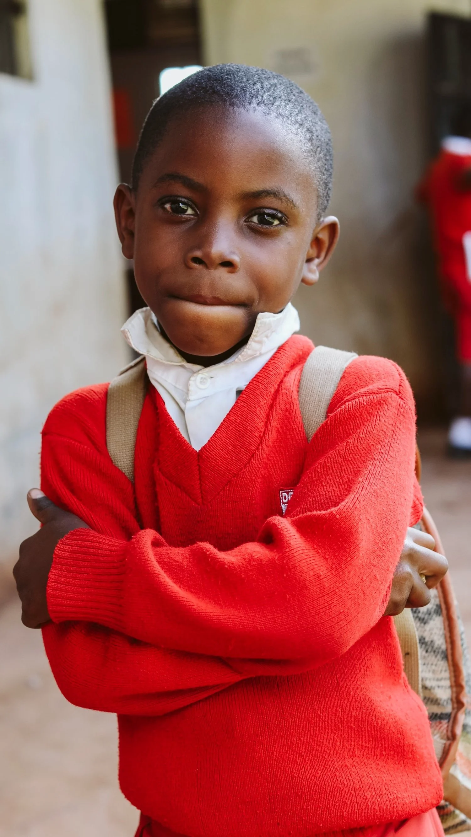 A young boy wearing a red sweater and a white shirt with a backpack over his shoulders, standing with his arms crossed inside a building or classroom.