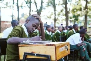 A classroom filled with students, mostly boys, sitting at desks outdoors, with some taking notes and appearing engaged in learning.
