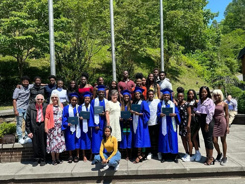 Group of graduate students in blue caps and gowns holding diplomas, standing outdoors with family and friends on a sunny day.