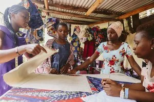 A group of women and girls gathered around a table, working on fabric or clothing, in an outdoor market or workshop setting.
