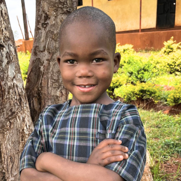 A young boy with a shaved head smiling and crossing his arms, standing outdoors next to a tree, with greenery and a building in the background.
