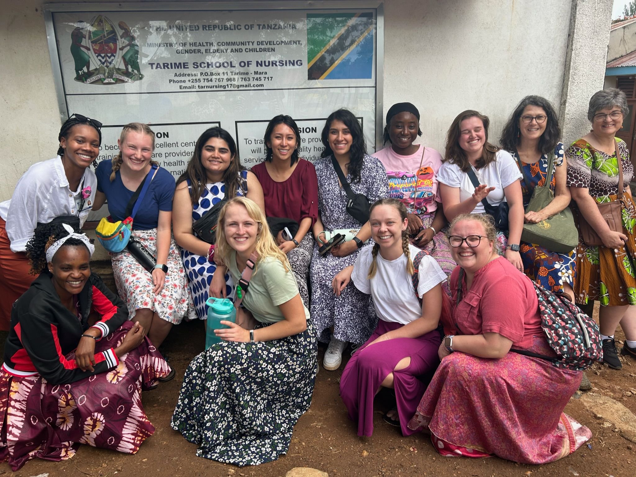 Group of young women and women standing and sitting in front of a sign for Tarime School of Nursing in Tanzania, smiling and posing for a photo.