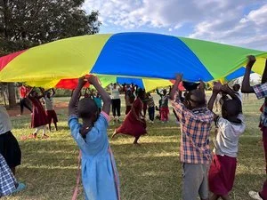 Children playing with a colorful parachute outdoors, with trees and a partly cloudy sky in the background.