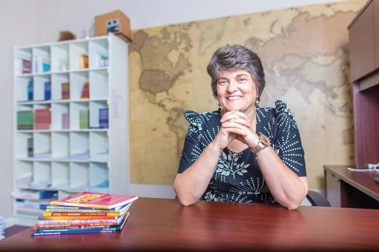 A woman with short gray hair and a patterned blue dress smiling at a desk with stacked colorful books, in an office with a large world map on the wall behind her.