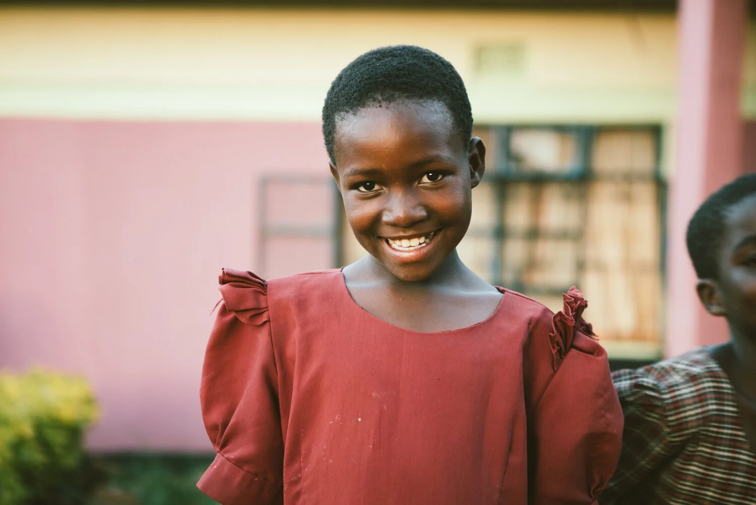 A young girl with dark skin, smiling, wearing a maroon dress with ruffled sleeves, outdoors near a pink building with a window and some greenery.
