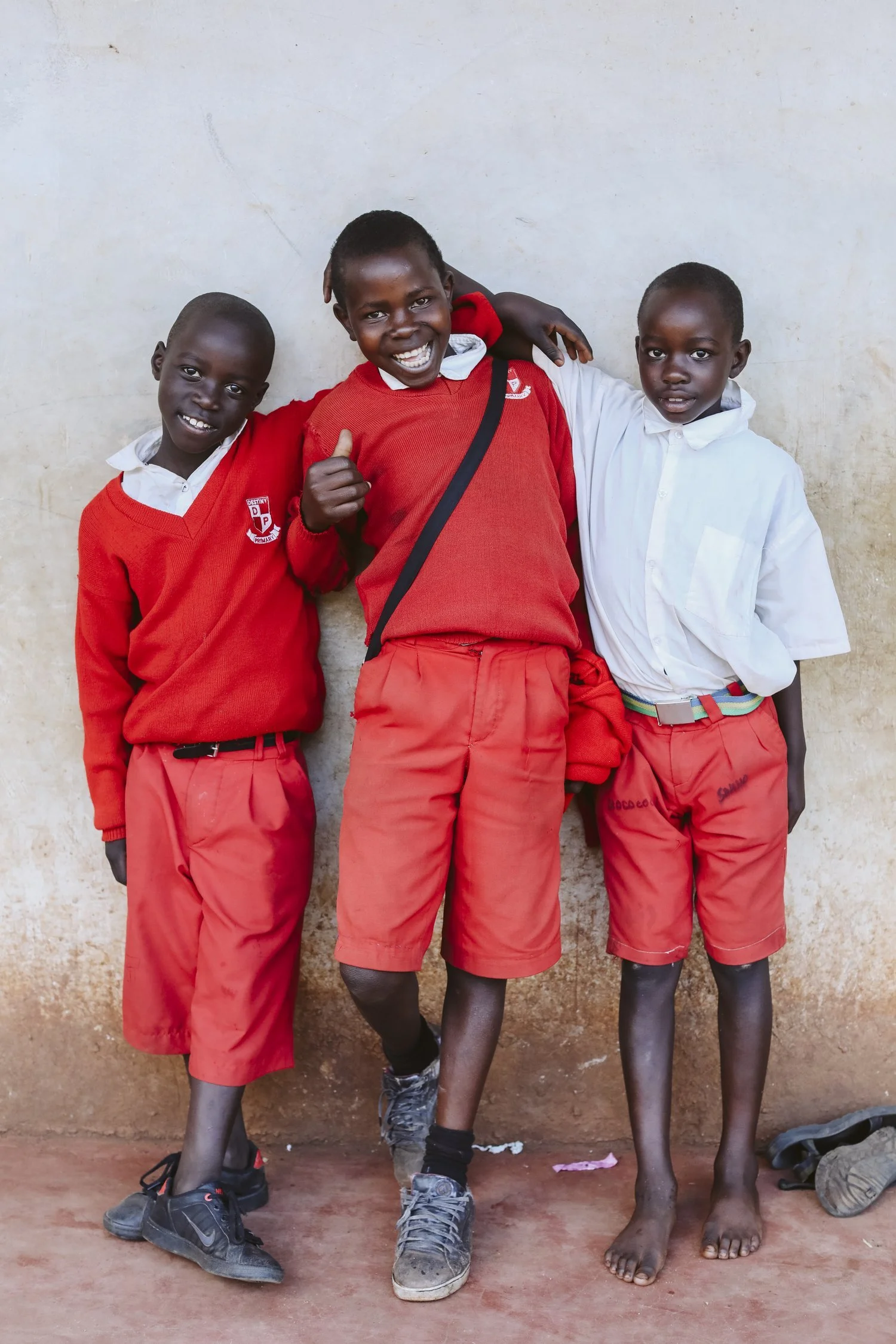 Three young boys in school uniforms smiling and standing against a wall, with one giving a thumbs-up.