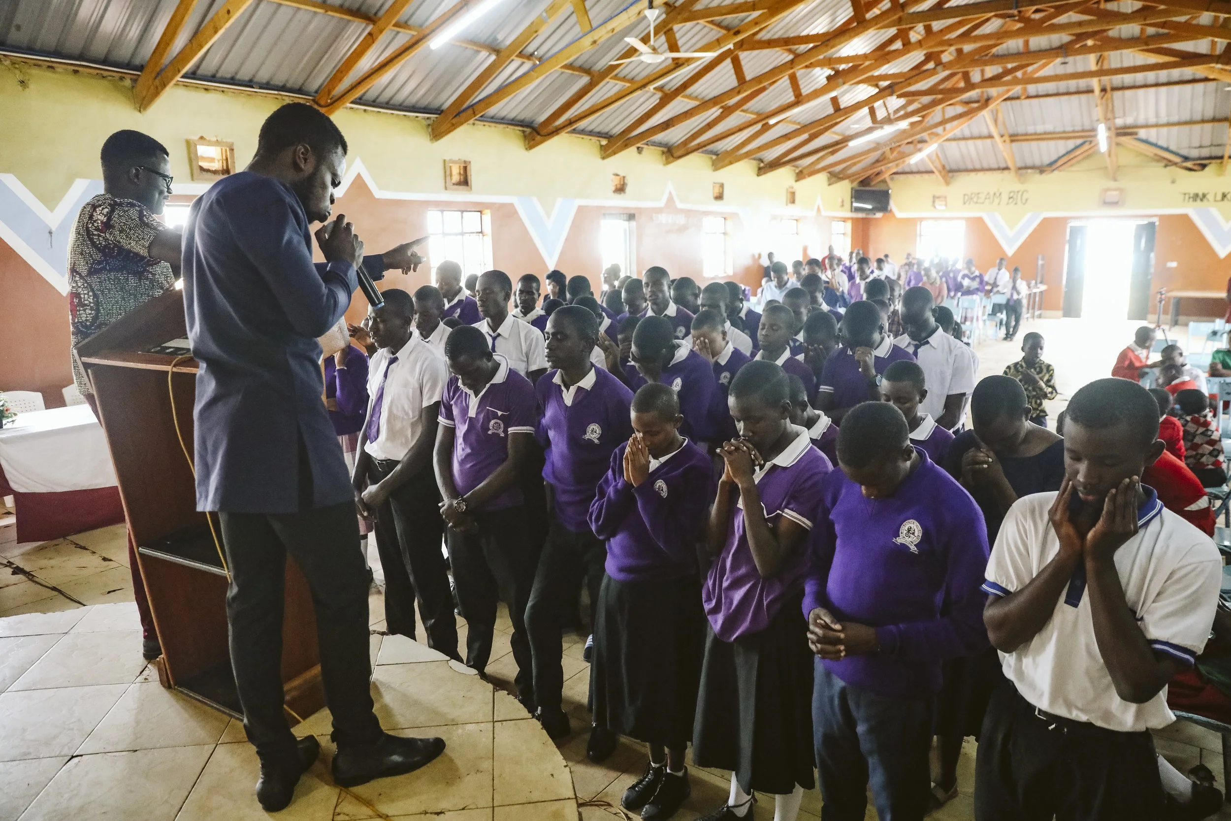 Students standing with hands clasped, praying during a church or prayer service in a large hall, with a man speaking at a podium.