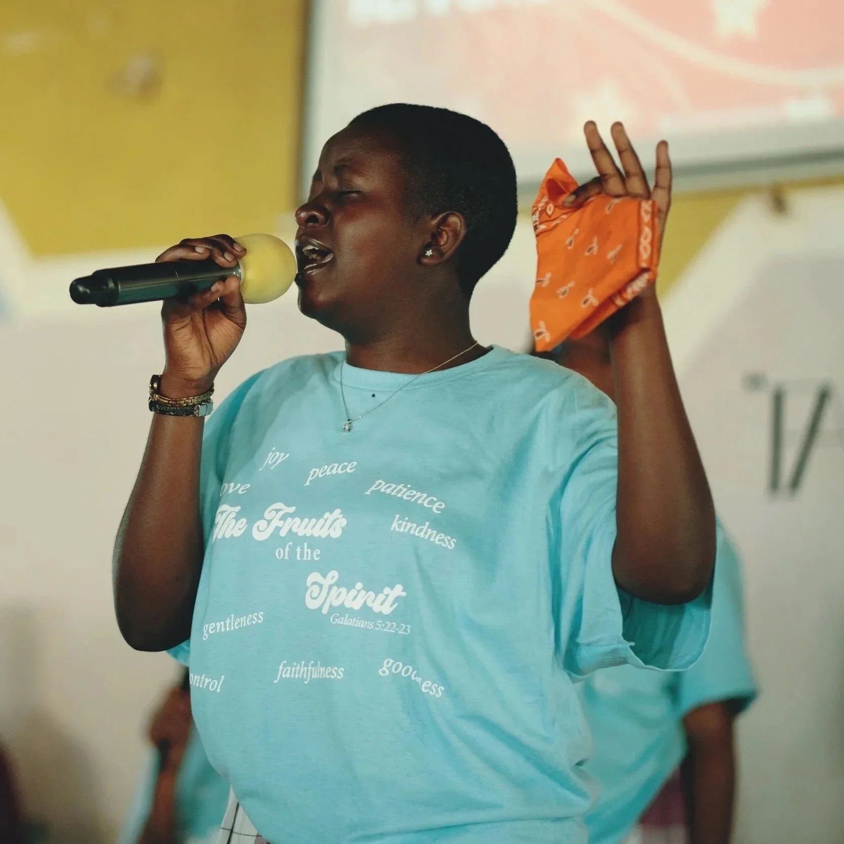A woman in a yellow jacket and red skirt instructs a group of children outdoors. The children, dressed in various colored shirts and skirts or shorts, appear engaged and attentive, with some raising their hands or gesturing.