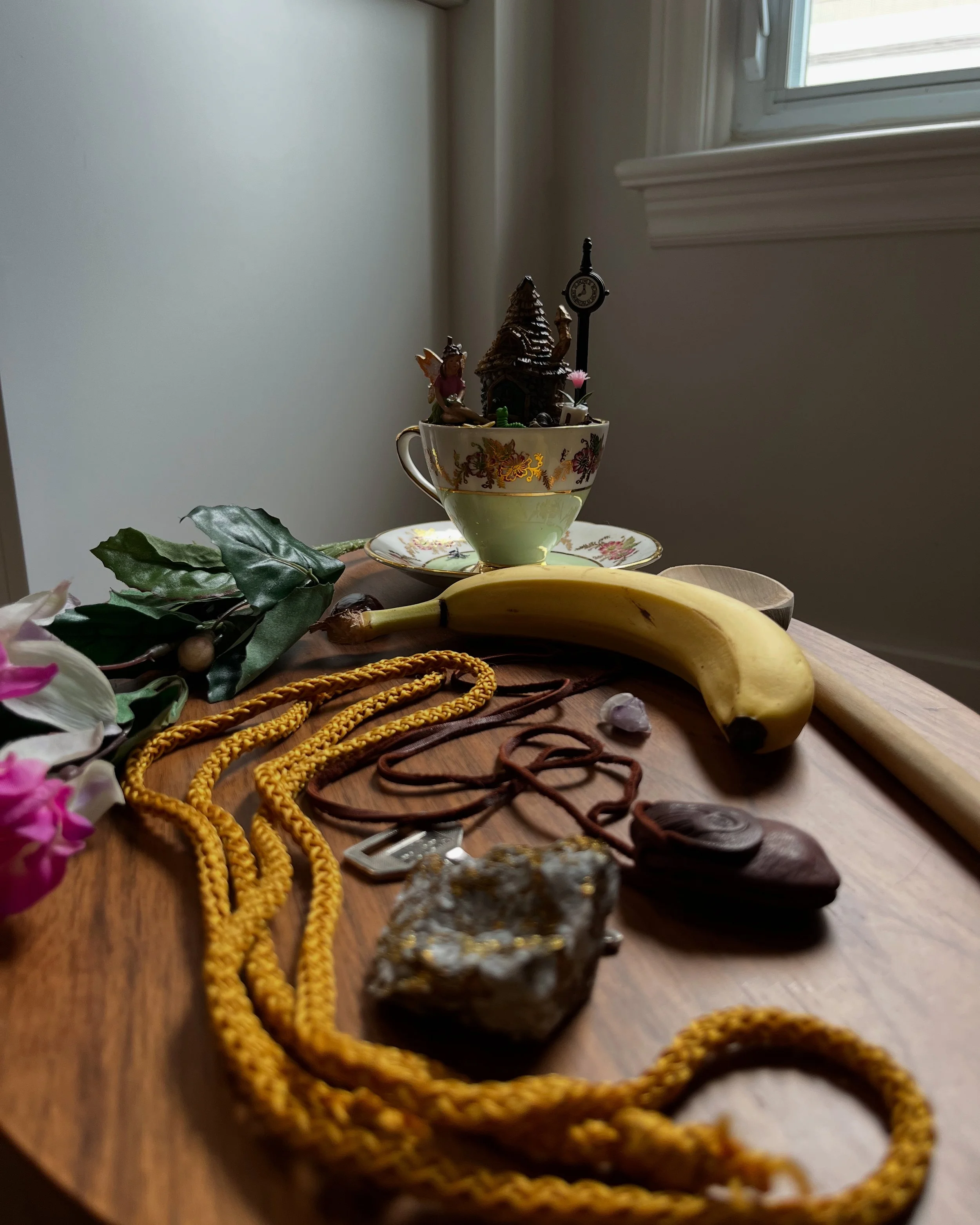 Collection of miscellaneous objects on a wooden table, including a yellow banana, a cup with miniature figurines and a castle, jewelry, a rock, a wooden spoon, a pink flower, and leafy greenery, with a window in the background.