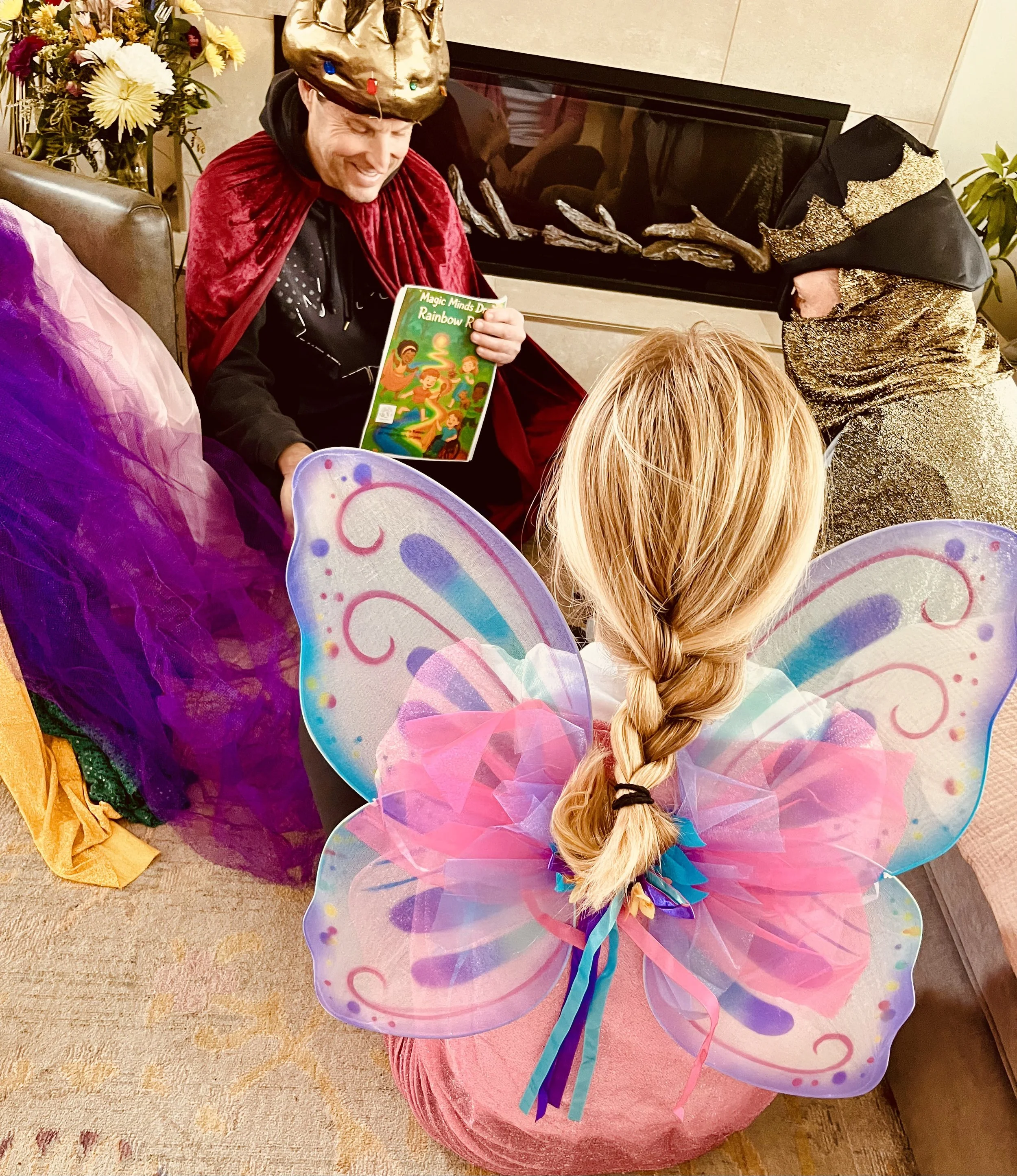A person dressed as a king with a crown and red cape reading a colorful book to a girl in fairy costume with pink and purple fairy wings and braid, sitting in a cozy room with a fireplace and flowers.