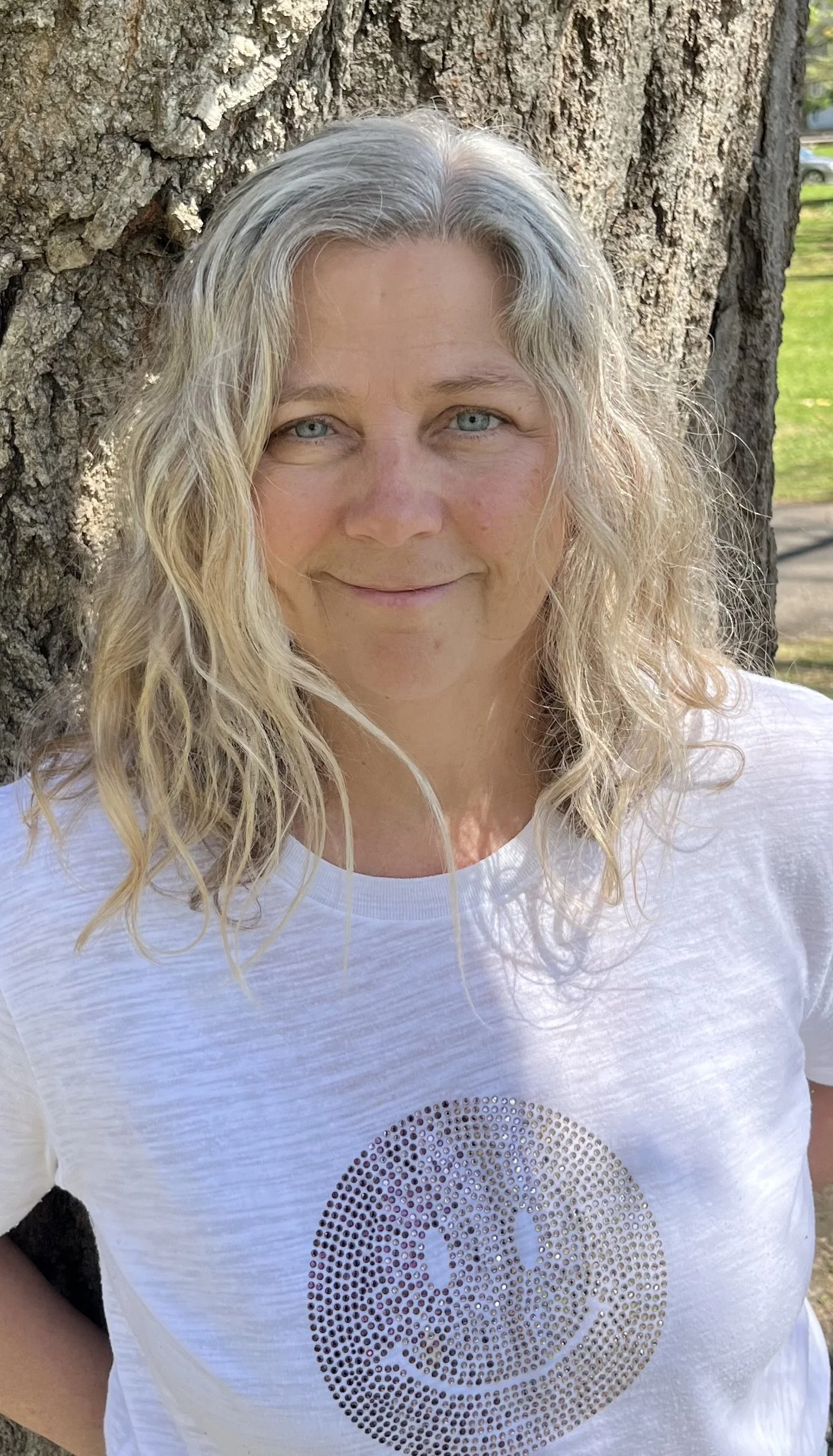 A woman with curly blonde hair and blue eyes smiling outdoors, leaning against a large tree trunk.
