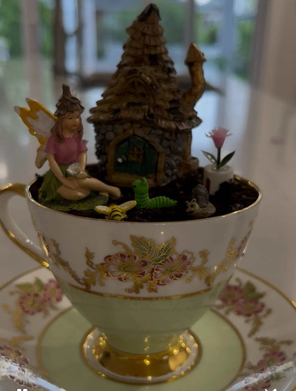 Decorative teacup with fairy garden miniature scene including a fairy figurine, a small house, a pink flower, a green caterpillar, a bee, and a hedgehog, all on soil-like material.