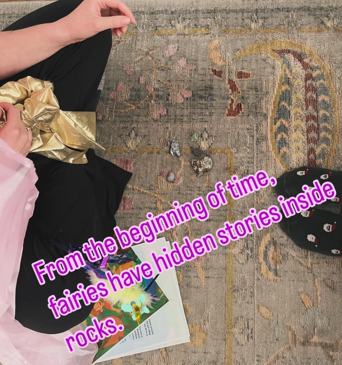 View of a colorful patterned rug with fairy stones and rocks arranged on it. Part of a person's legs in black pants, another person's hand holding a gold gift bag, a children's book with a fairy illustration, and a pair of feet in black socks with small white and red designs. Text overlay in purple says: "From the beginning of time, fairies have hidden stories inside rocks."