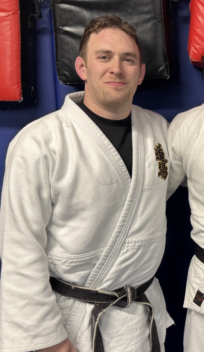 A man in a white martial arts gi with black belt, smiling, standing against a blue wall with martial arts equipment, including red and black pads.