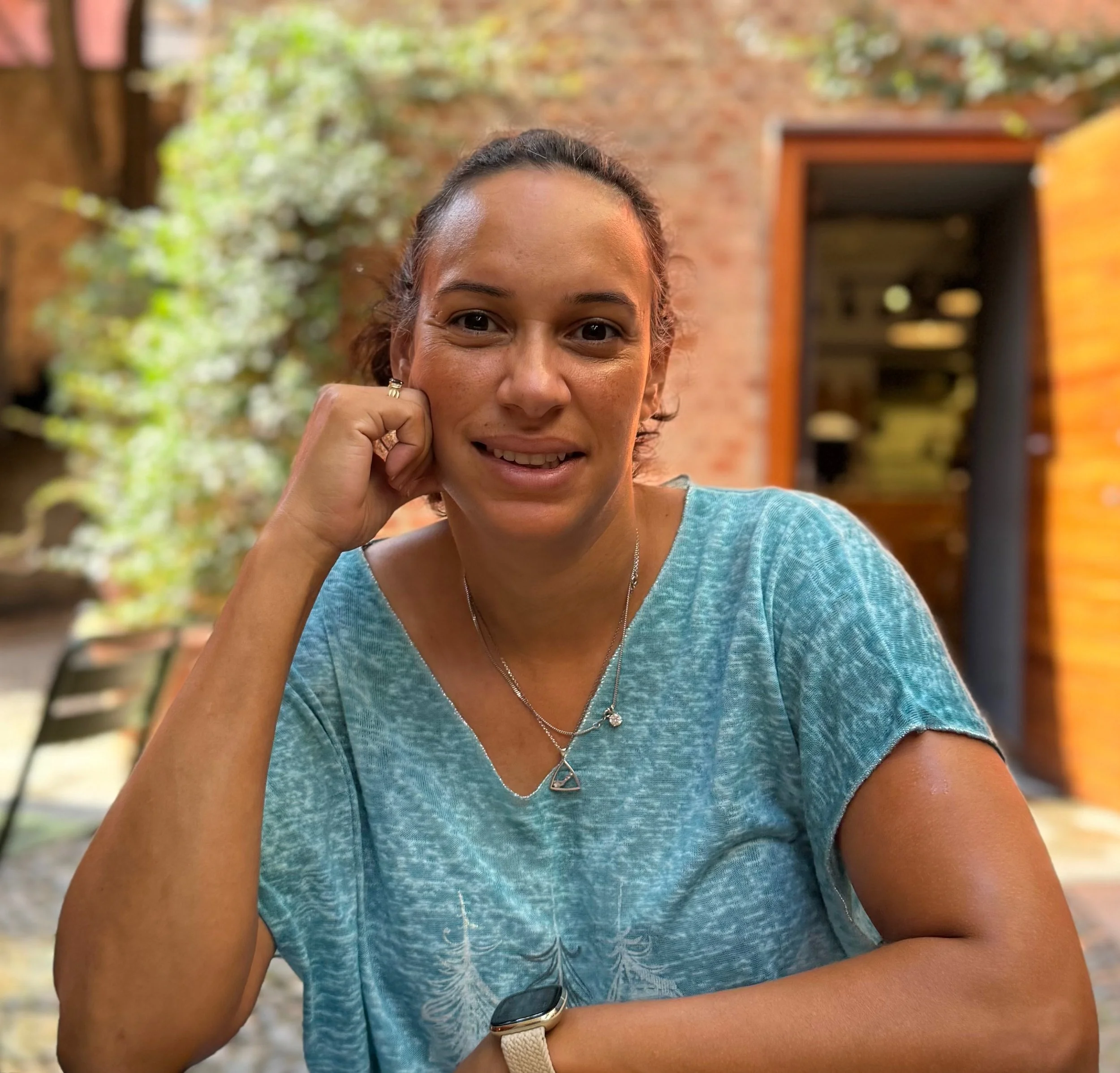 A person in a blue shirt sitting in an outdoor setting by a brick wall, smiling with their hand resting on their chin.