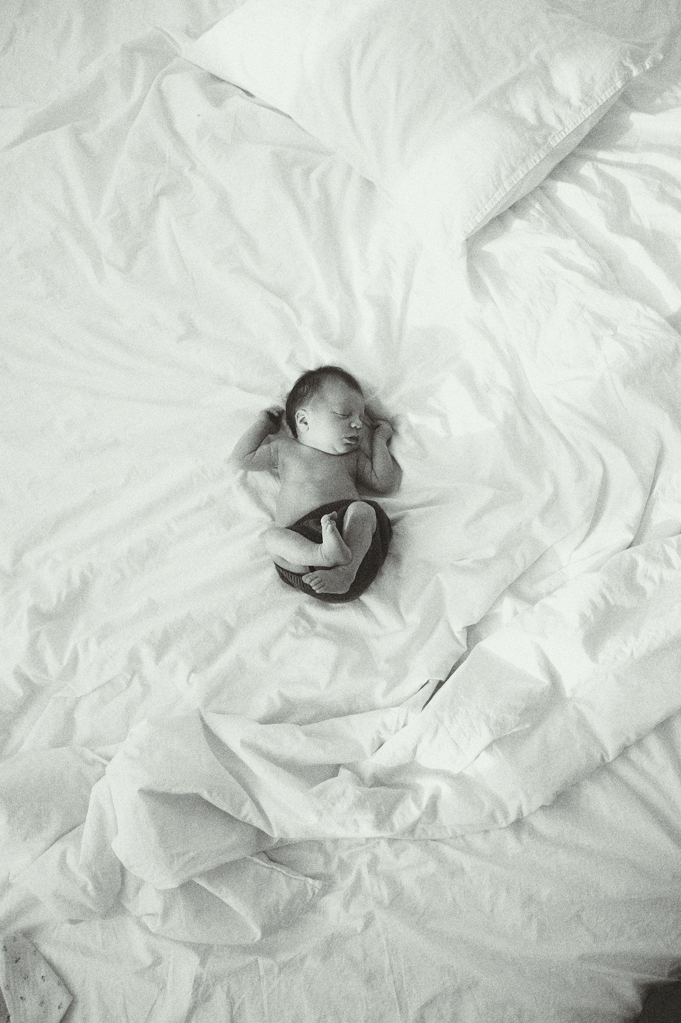 Black and white photo of a newborn baby sleeping on a bed with white blankets and pillow.