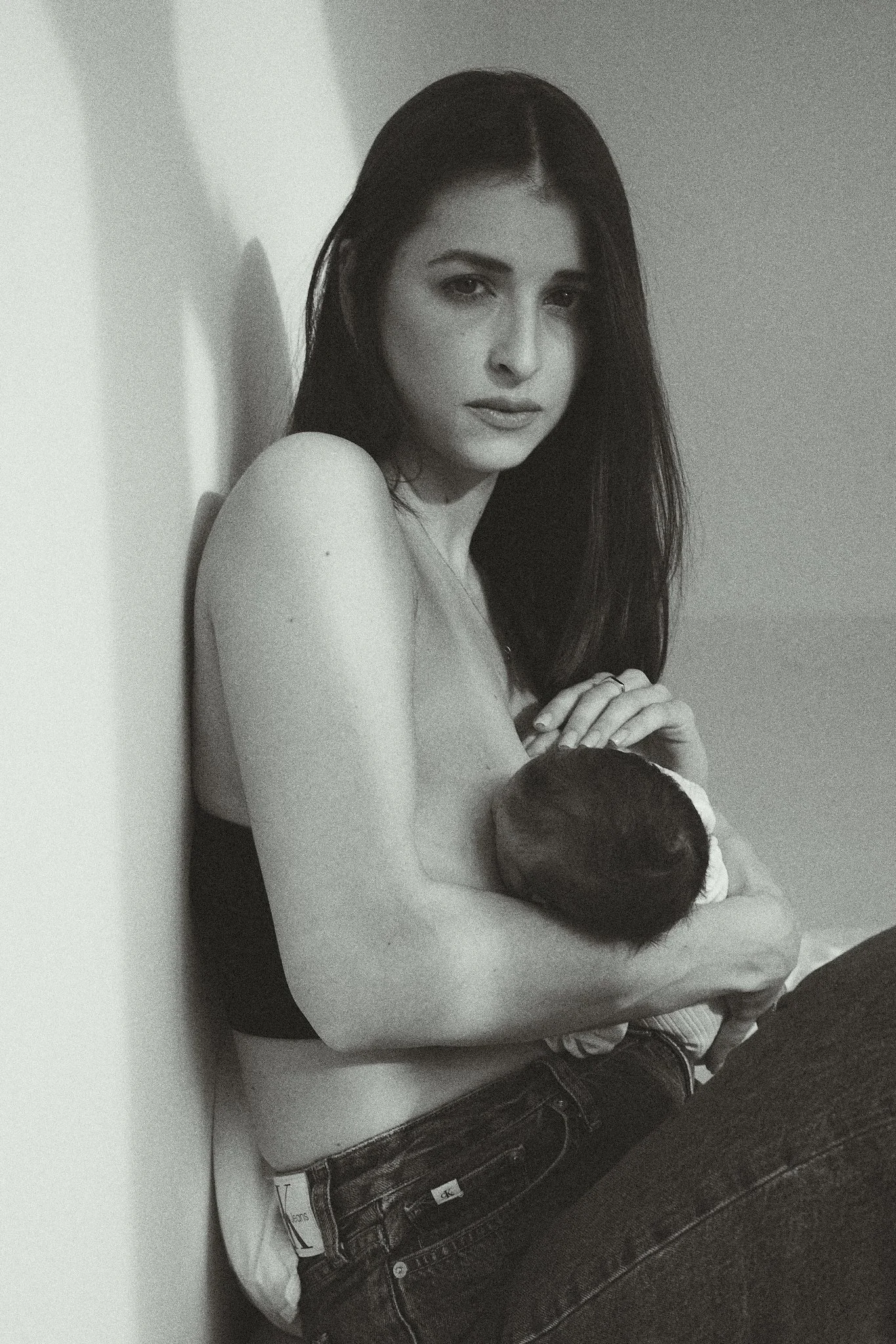 Woman breastfeeding a baby, sitting against a wall, in a black and white photo.