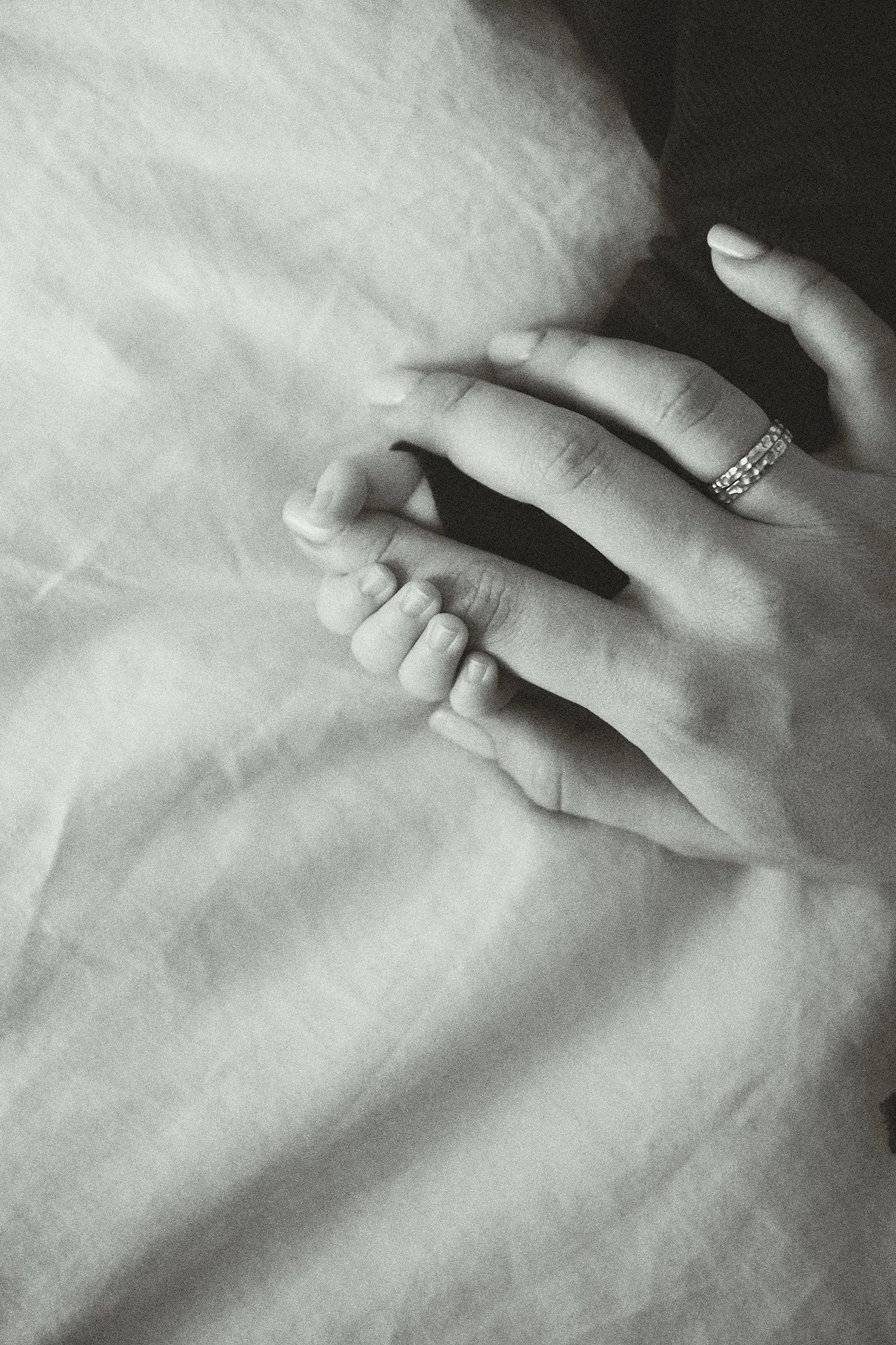 Close-up of two hands holding each other on a white sheet, one wearing a ring.