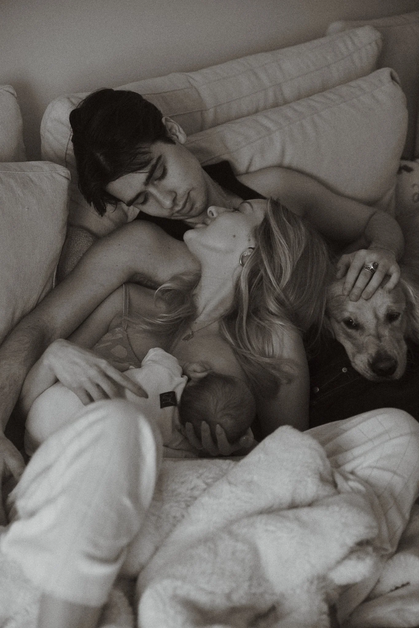 A black and white photo of a couple lying on a couch, holding a baby and a dog.
