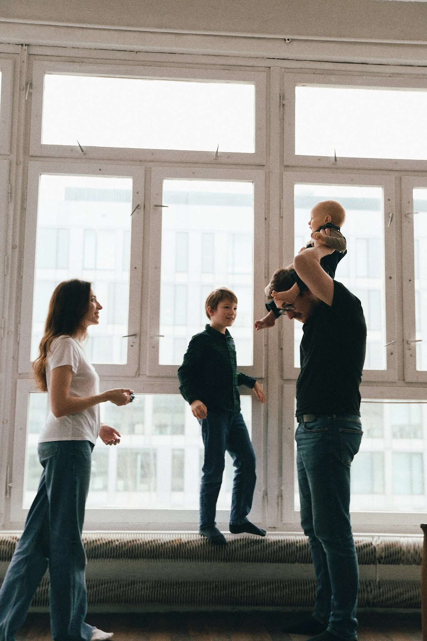 Family playing indoors, father holding baby, boy standing on radiator, mother watching, large windows and buildings in background.