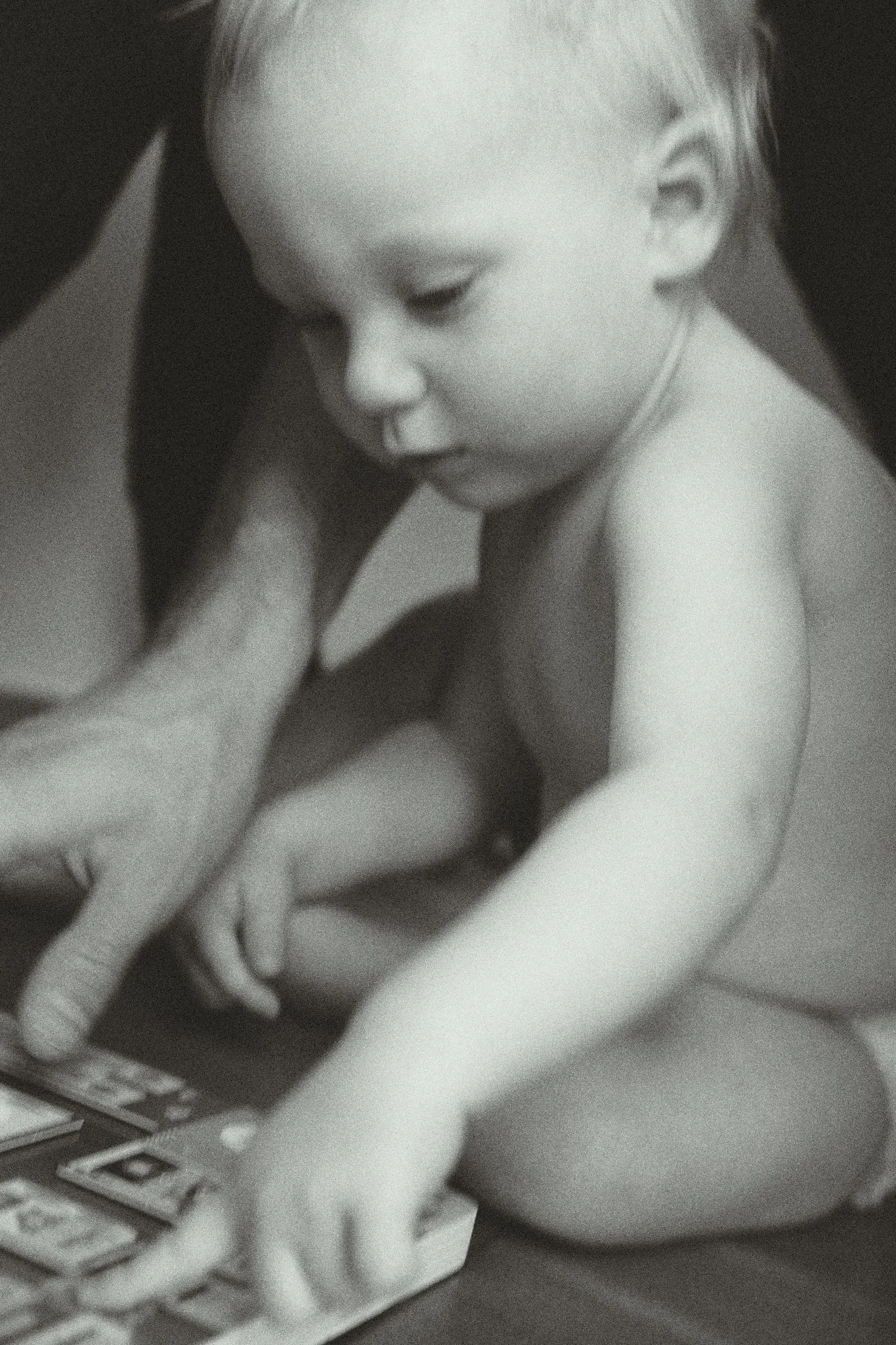 Black and white photo of a baby playing with cards on the floor, with an adult hand assisting.