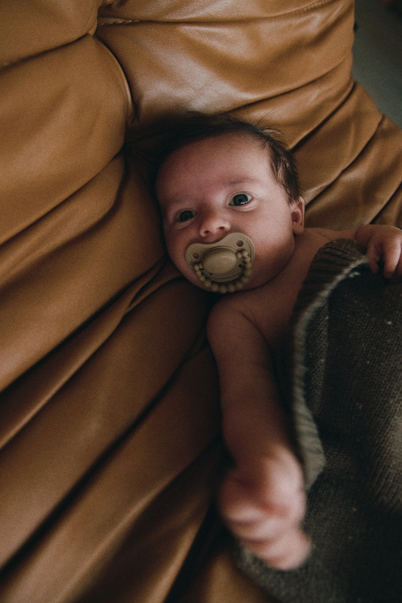 A baby lying on a brown leather couch with a pacifier in its mouth, partially covered by a knitted blanket.