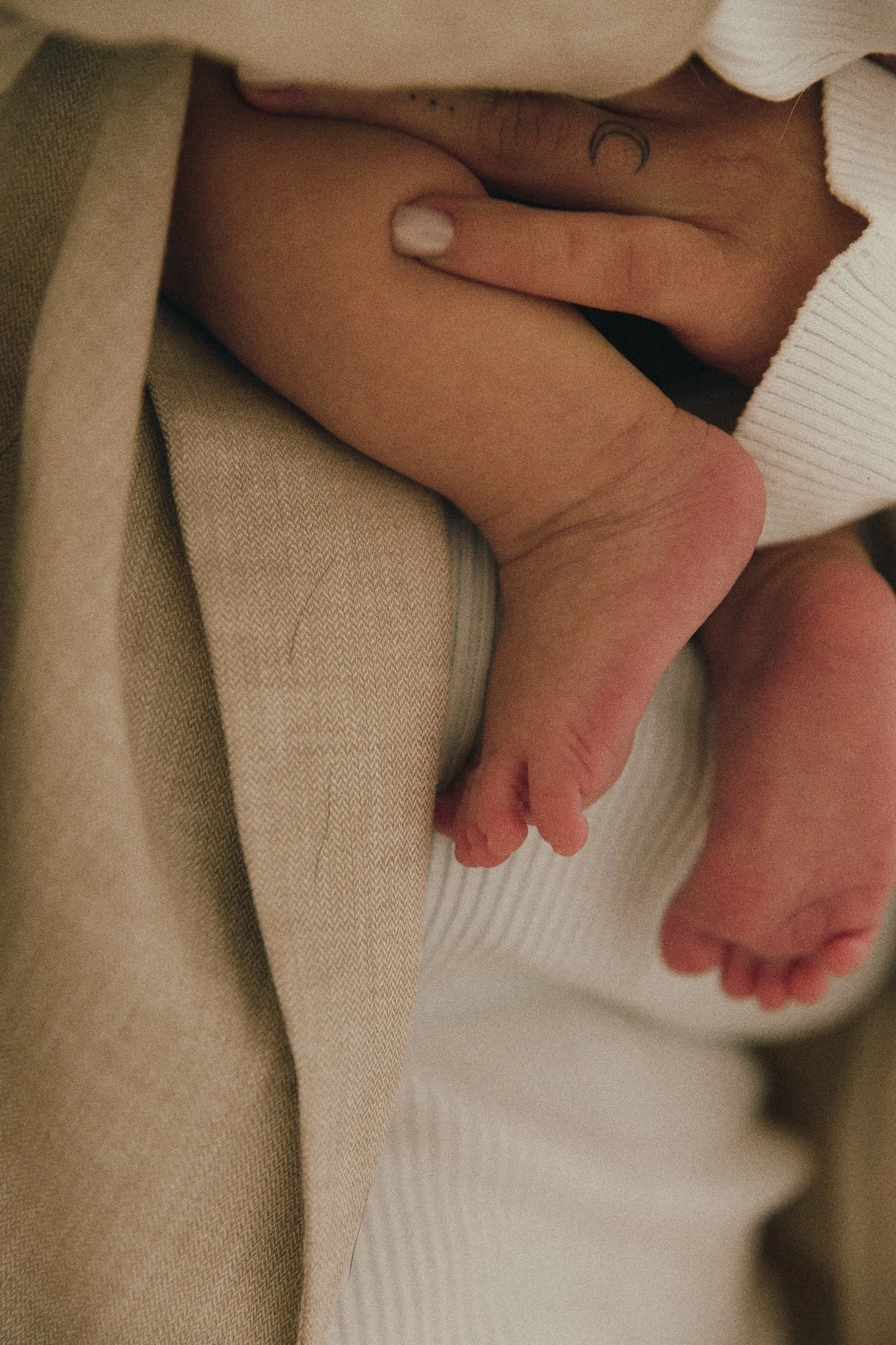 Close-up of a person's hand gently holding a baby's legs and feet, with a tattoo visible on the hand.
