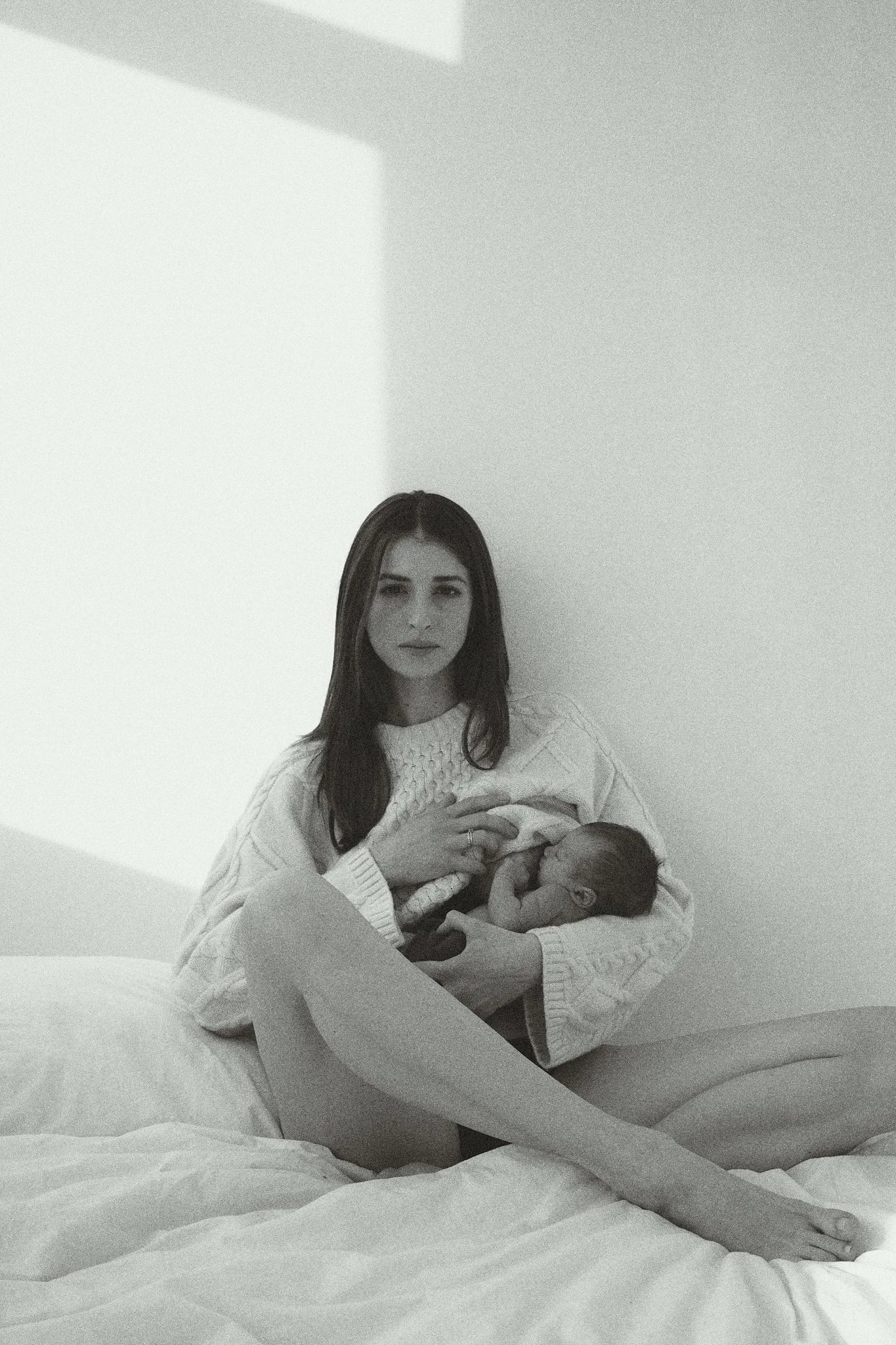 Woman in a sweater holding a baby while sitting on a bed in a softly lit room.