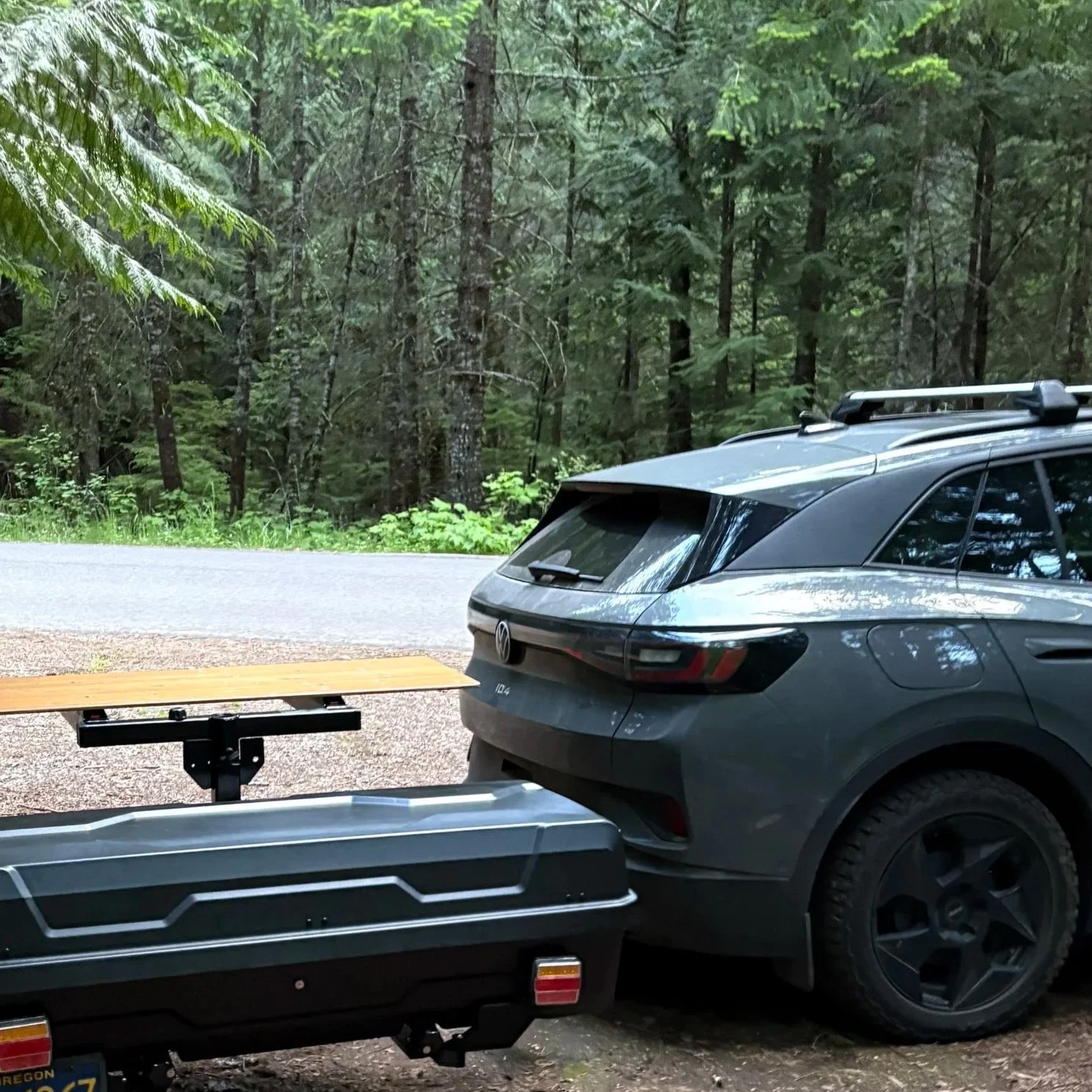 A gray Volkswagen ID4 electric SUV parked outdoors in a wooded area, next to a trailer with a wooden platform, in a natural, forested environment.