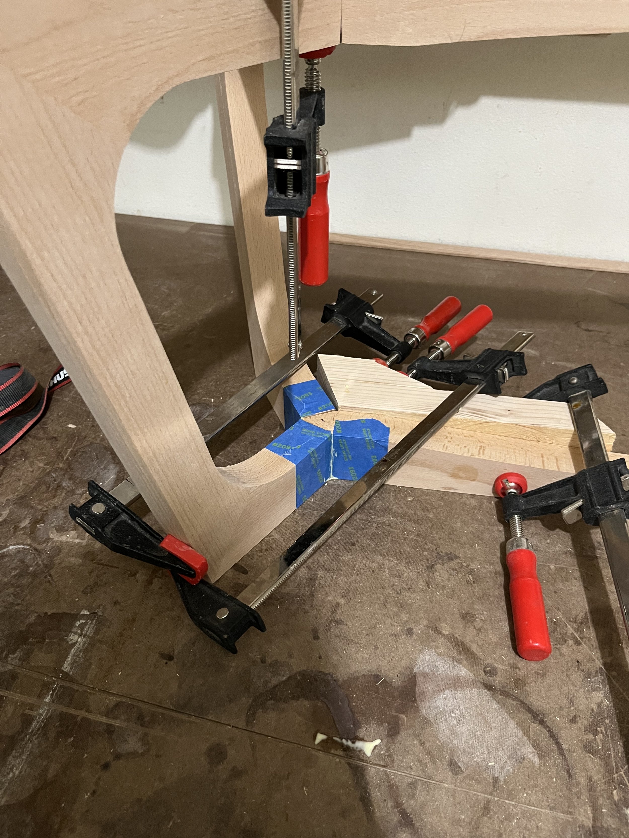 A woodworking project with a partially assembled wooden chair or table. Clamps and blue painter's tape are used to hold the pieces together while the glue dries.