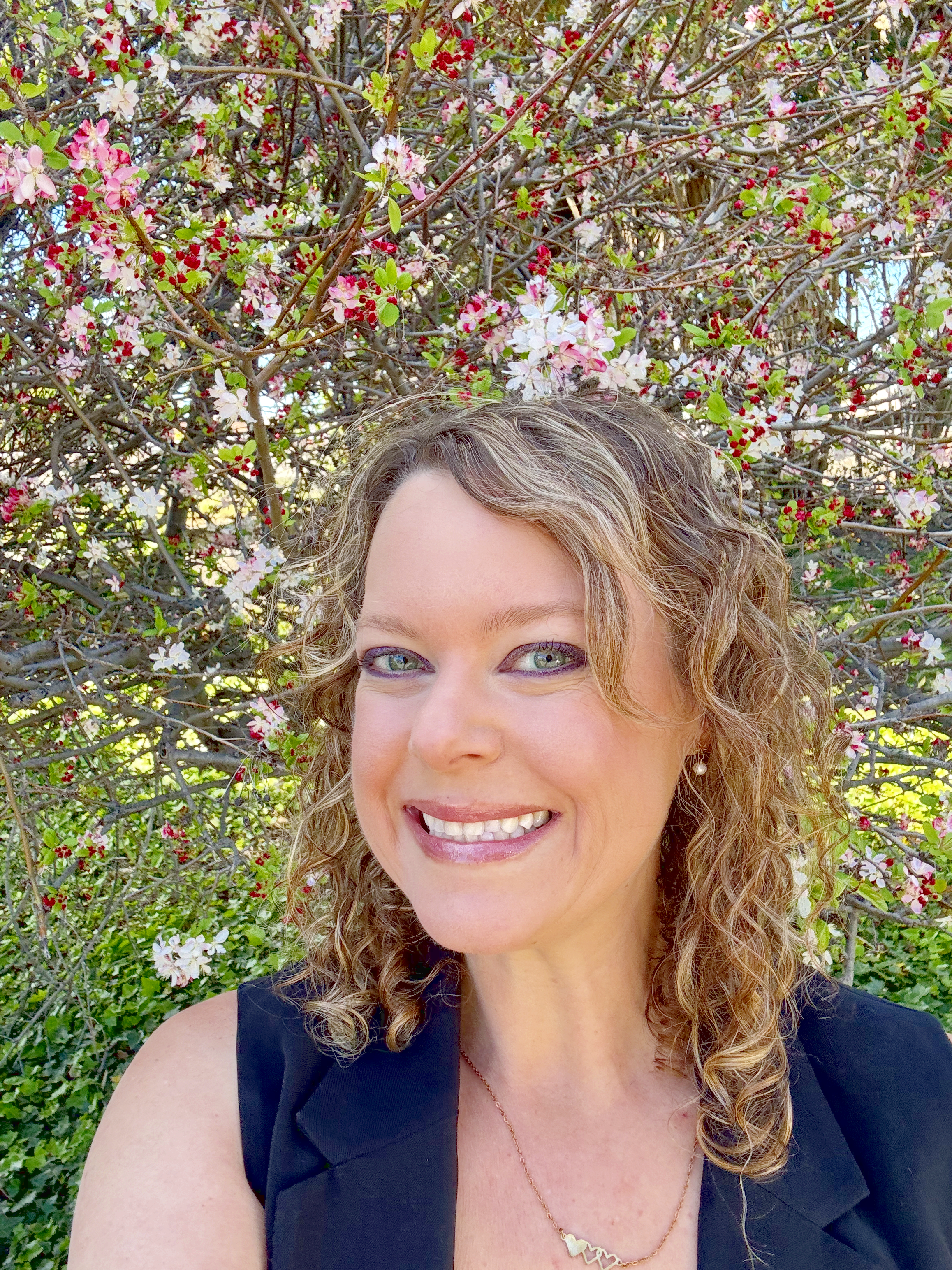 A woman with curly blonde hair smiling, standing outdoors in front of pink flowers and green leaves.