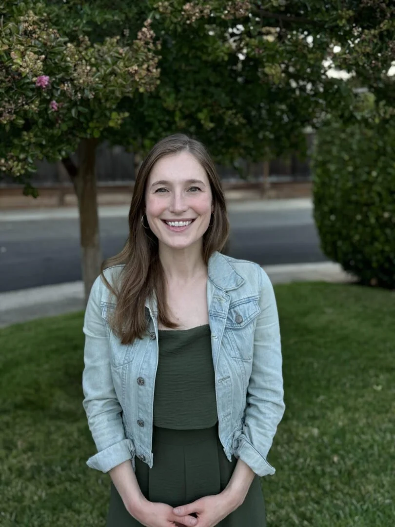 A young woman standing outdoors on a grassy area, smiling at the camera with trees and a street in the background.
