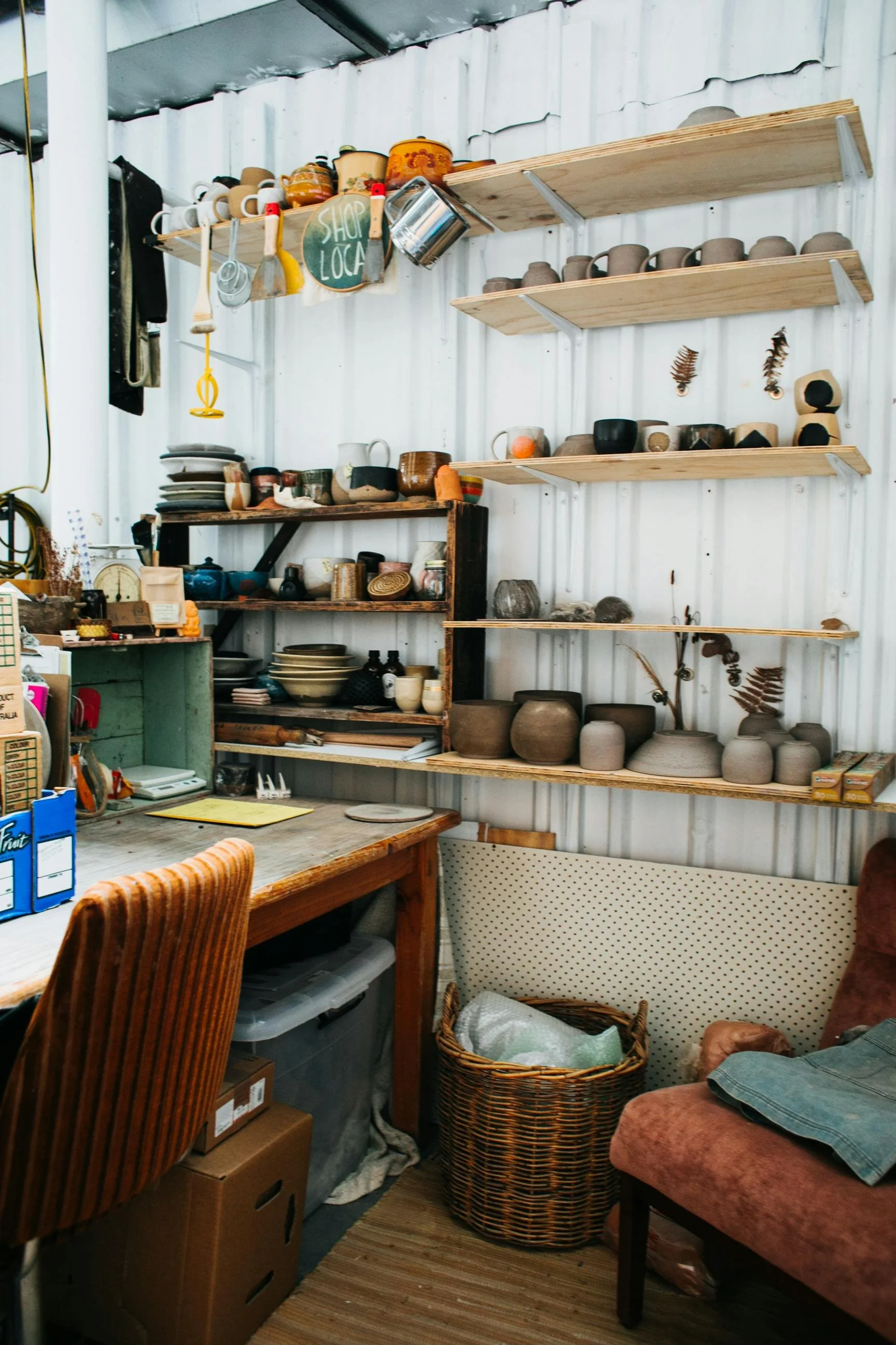Een interieur met houten planken en een verzameling keramische bekers, kommen en vazen op planken. Er staan ook decoratieve bladeren aan de muur en winkelschappen met keramiek en andere voorwerpen.