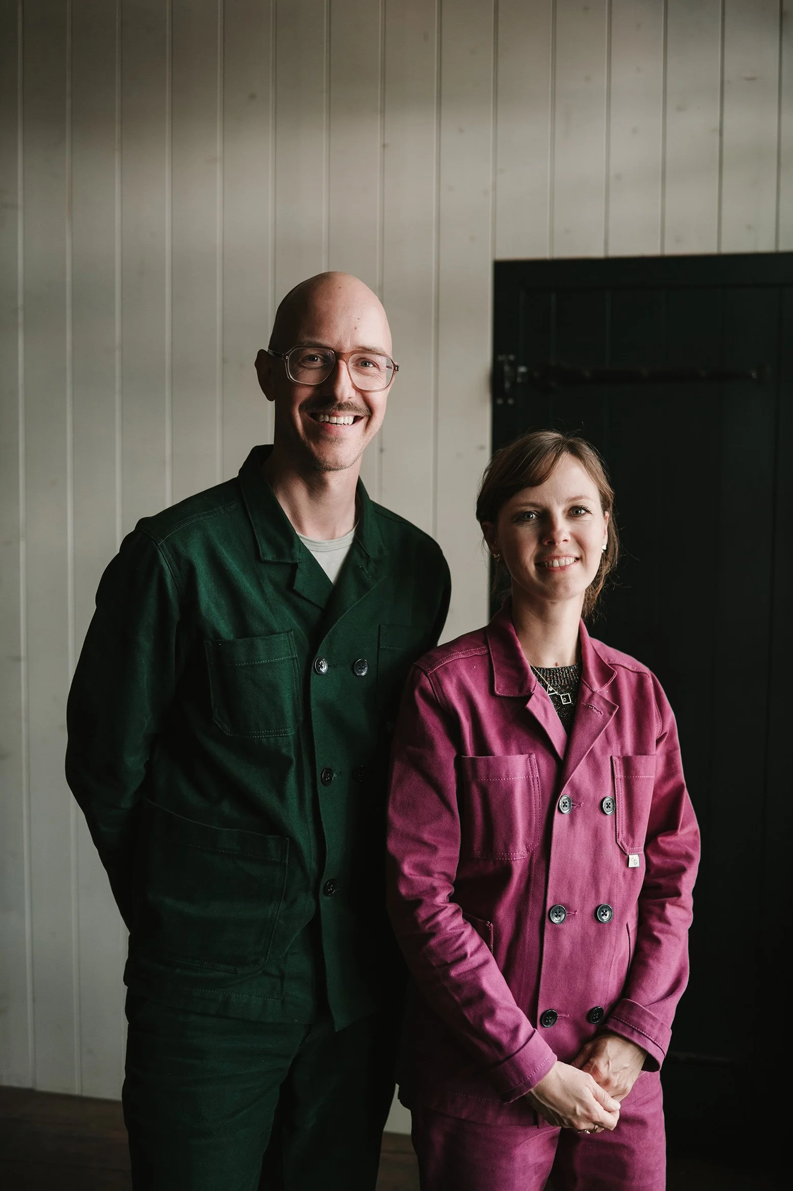 Twee mensen, een man en een vrouw, staan samen voor een houten paneelwand. De man draagt een groene jas en heeft een bril, de vrouw draagt een roze jas en heeft bruin haar.