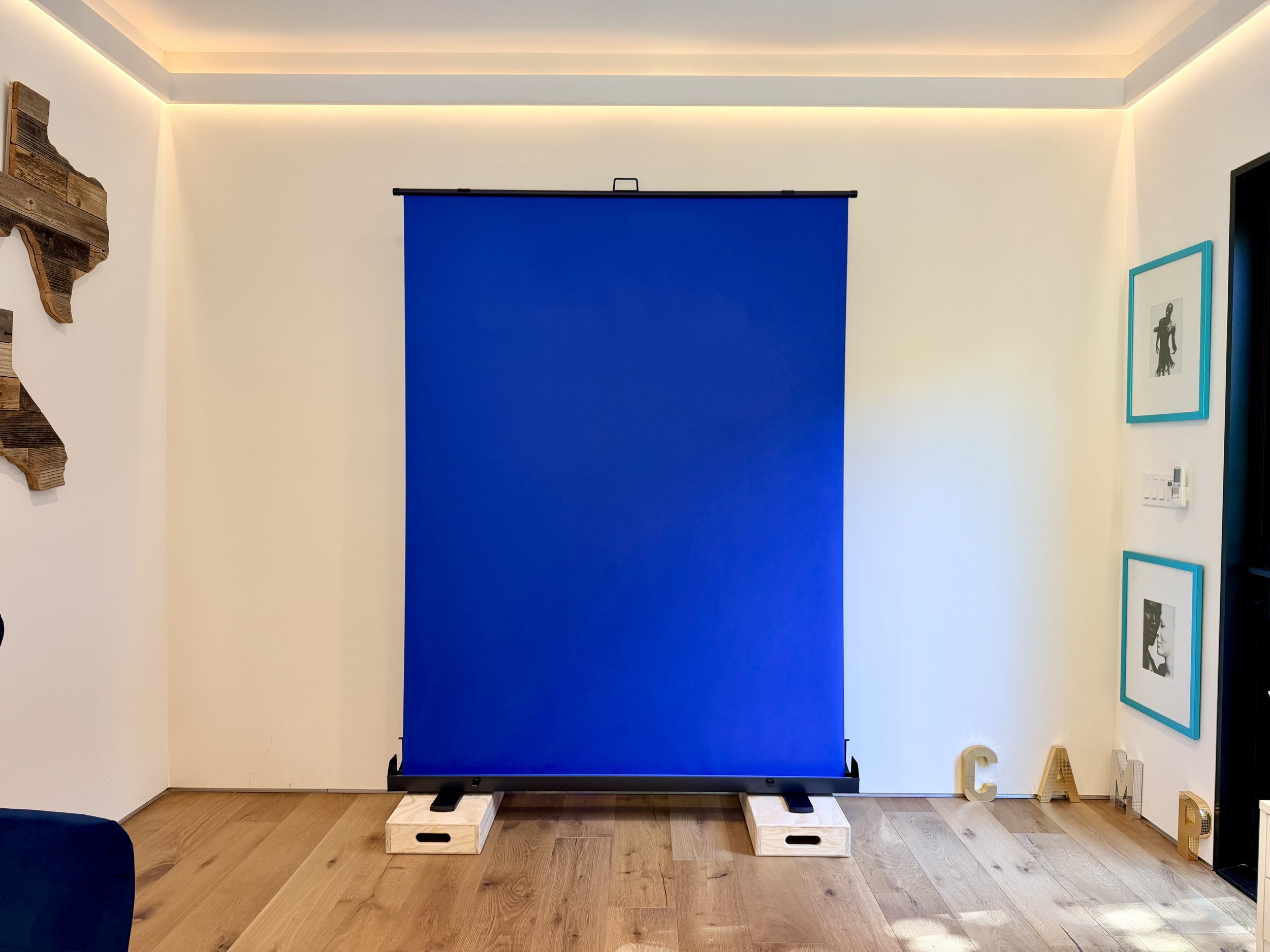A blue photography backdrop on a stand in a room with white walls and framed pictures. Wooden blocks are at the base of the stand, and wooden letters spelling 'CALI' are on the floor nearby.