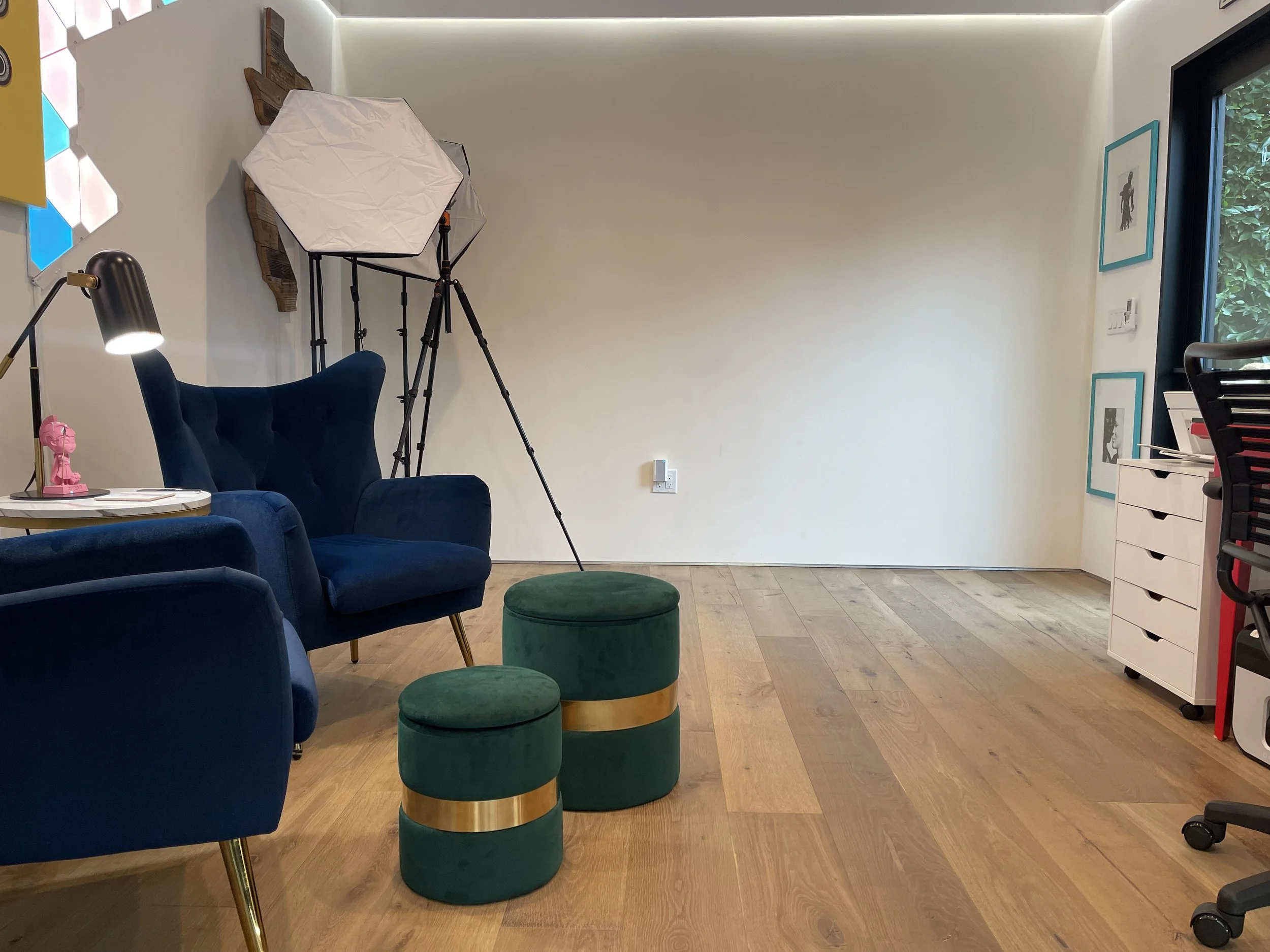A room with a blue tufted armchair and matching footstool, a green velvet stool with gold band, a white end table with a pink figurine and a black desk lamp, a white storage unit, and an art development on the wall with a large window showing greener