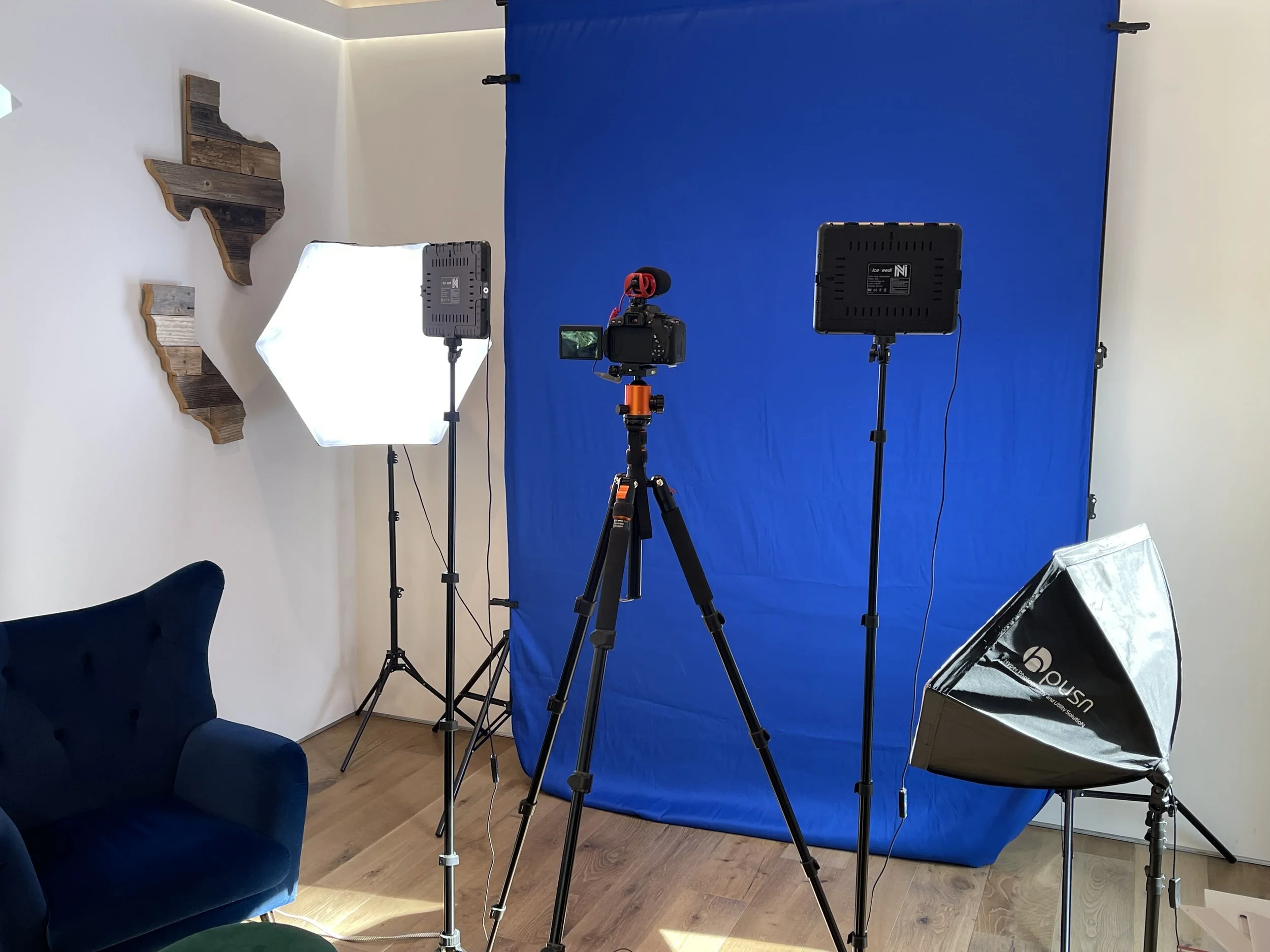 Interior of a video production studio with a blue backdrop, professional lighting equipment, camera on tripod, and a blue velvet chair, with wooden wall decor and hardwood flooring.