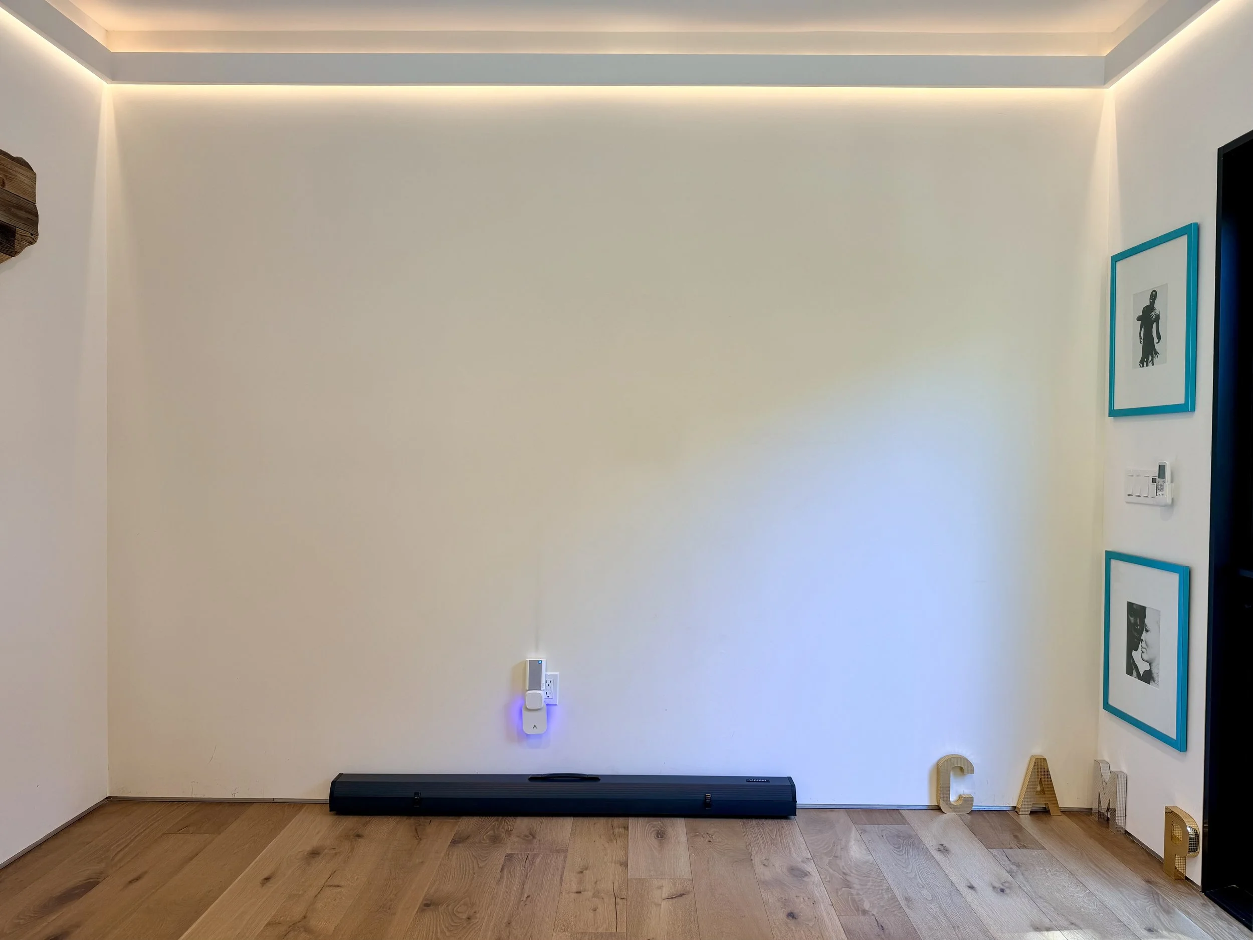 Empty living room corner with white wall, framed artwork, decorated letters spelling C A M, a heater, and a light switch.