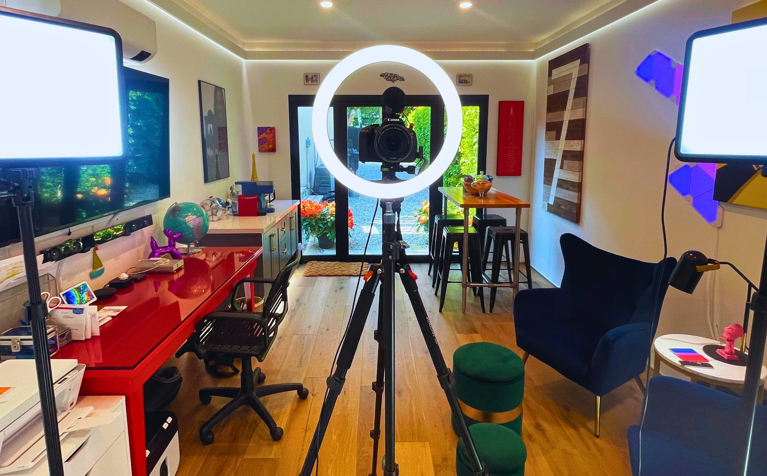 A room set up for filming or photography, with a Canon camera on a tripod surrounded by two computer monitors, a red desk, a black office chair, a small round table with pink and blue objects, a bar-height table with three stools, and various decorat